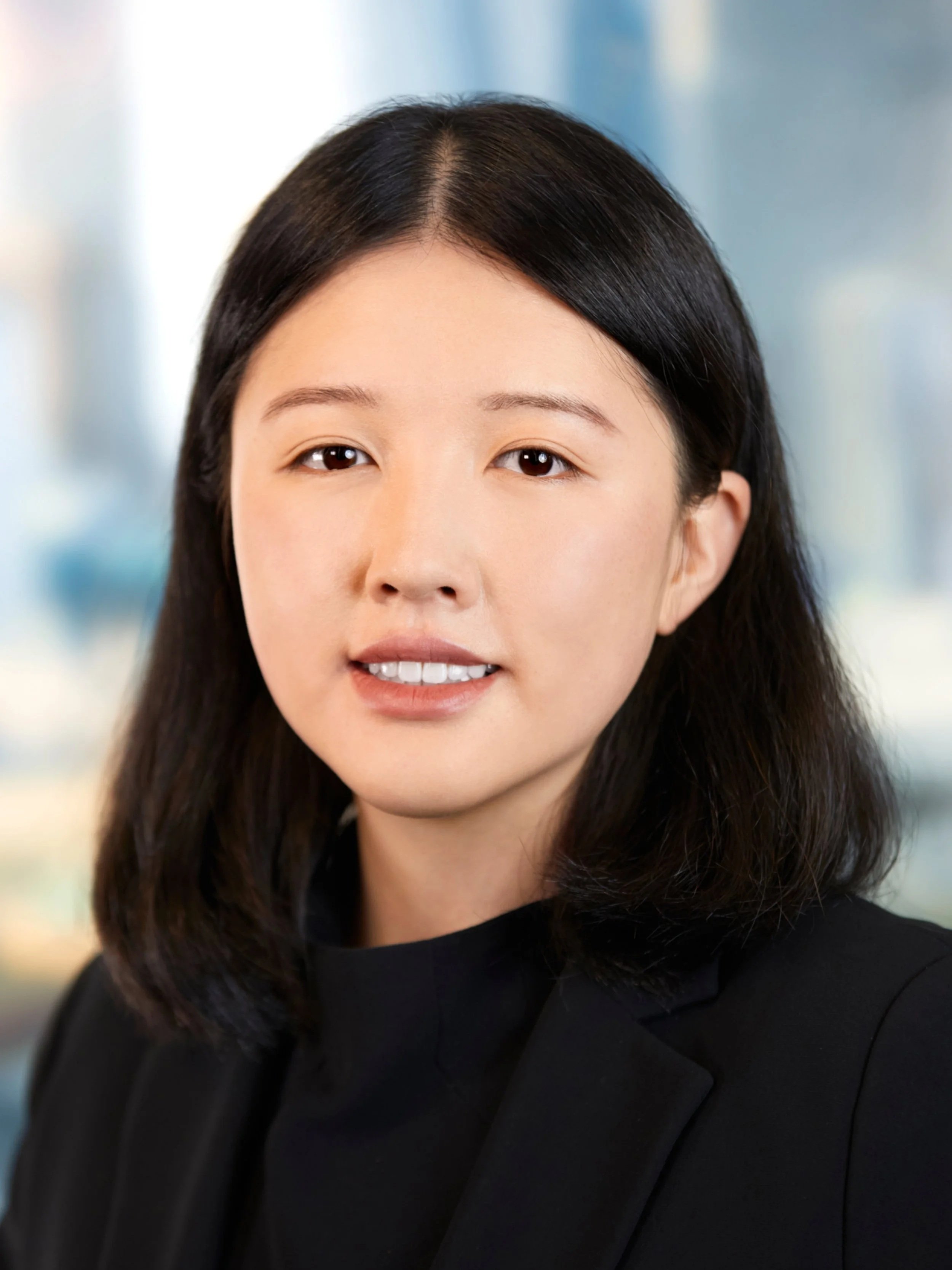 A professional portrait of a woman with shoulder-length black hair, wearing a black blazer and a black top, against a blurred modern office background.