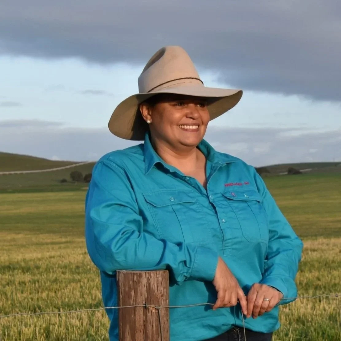 A woman wearing a wide-brimmed hat and a blue outdoor shirt standing by a wooden fence in a grassy field with rolling hills and a cloudy sky in the background.