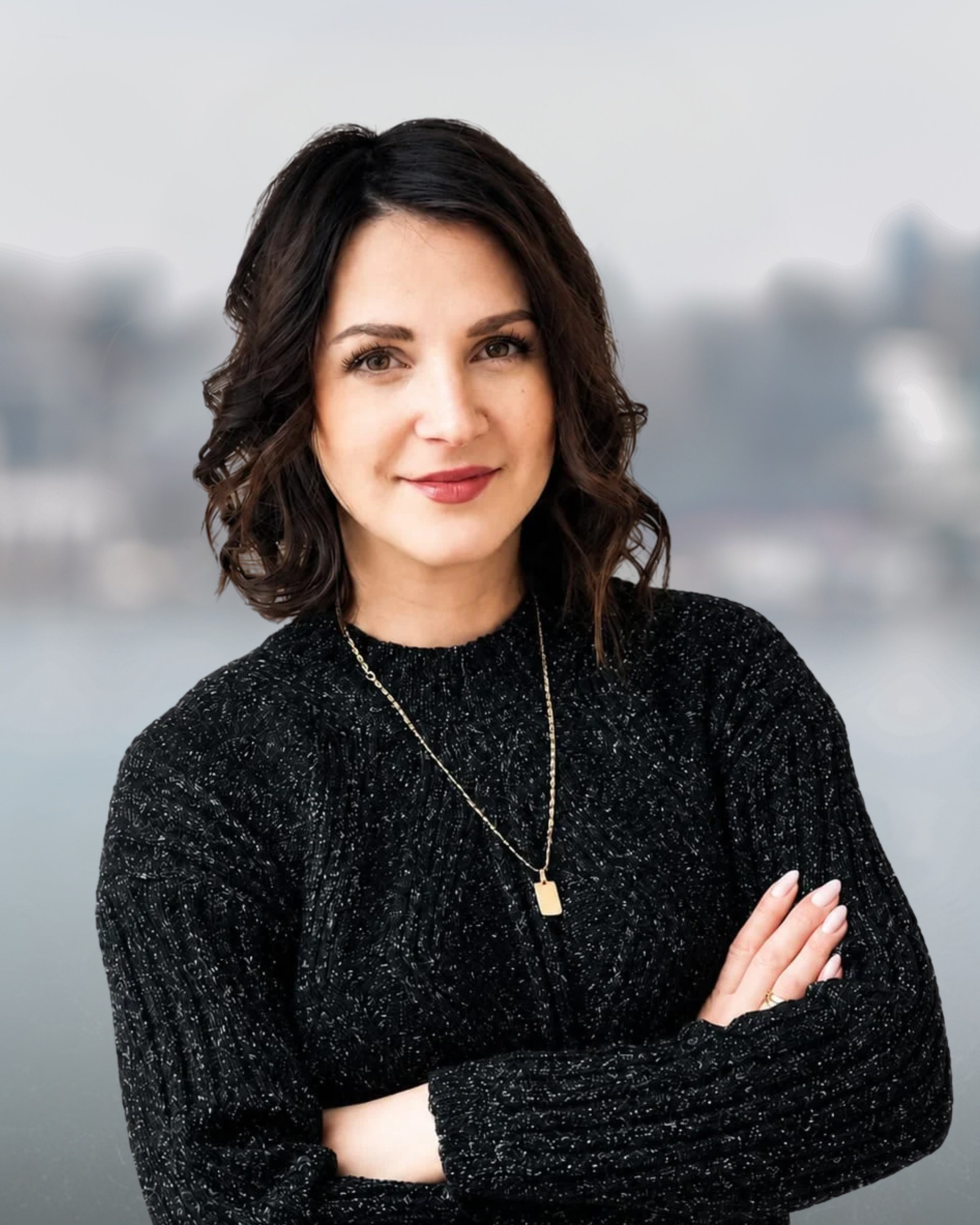 Portrait of a woman with long dark hair, wearing a navy blazer and a necklace, against a blurred outdoor background.