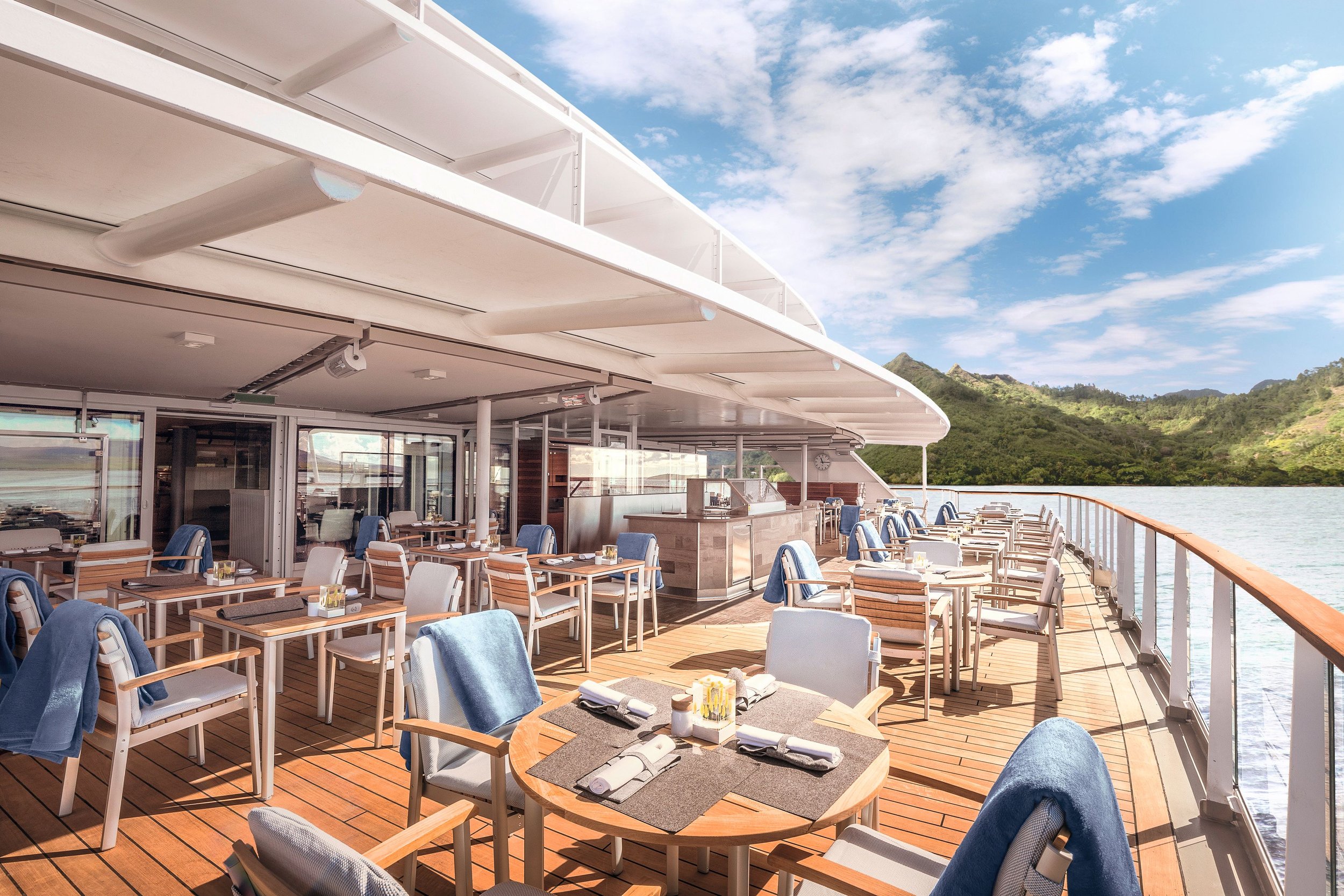 Outdoor dining area on a luxury cruise ship with set tables, chairs, and a view of the ocean and distant hills.