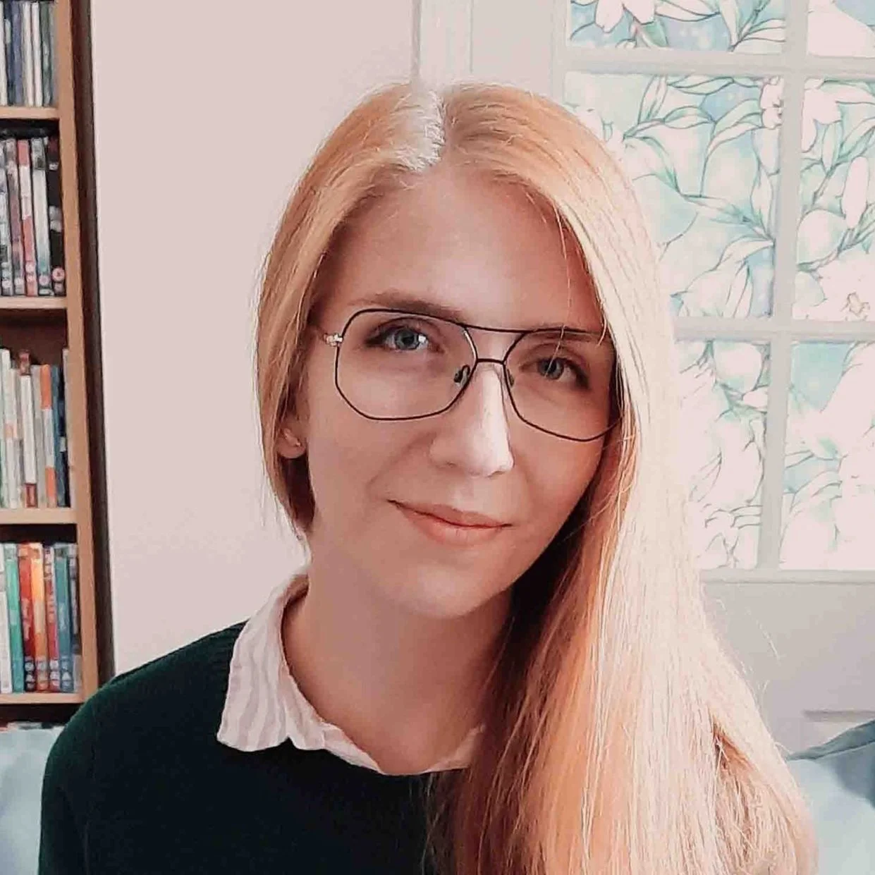 Person with long blonde hair and glasses smiling indoors, with a decorative glass window and a bookshelf in the background.