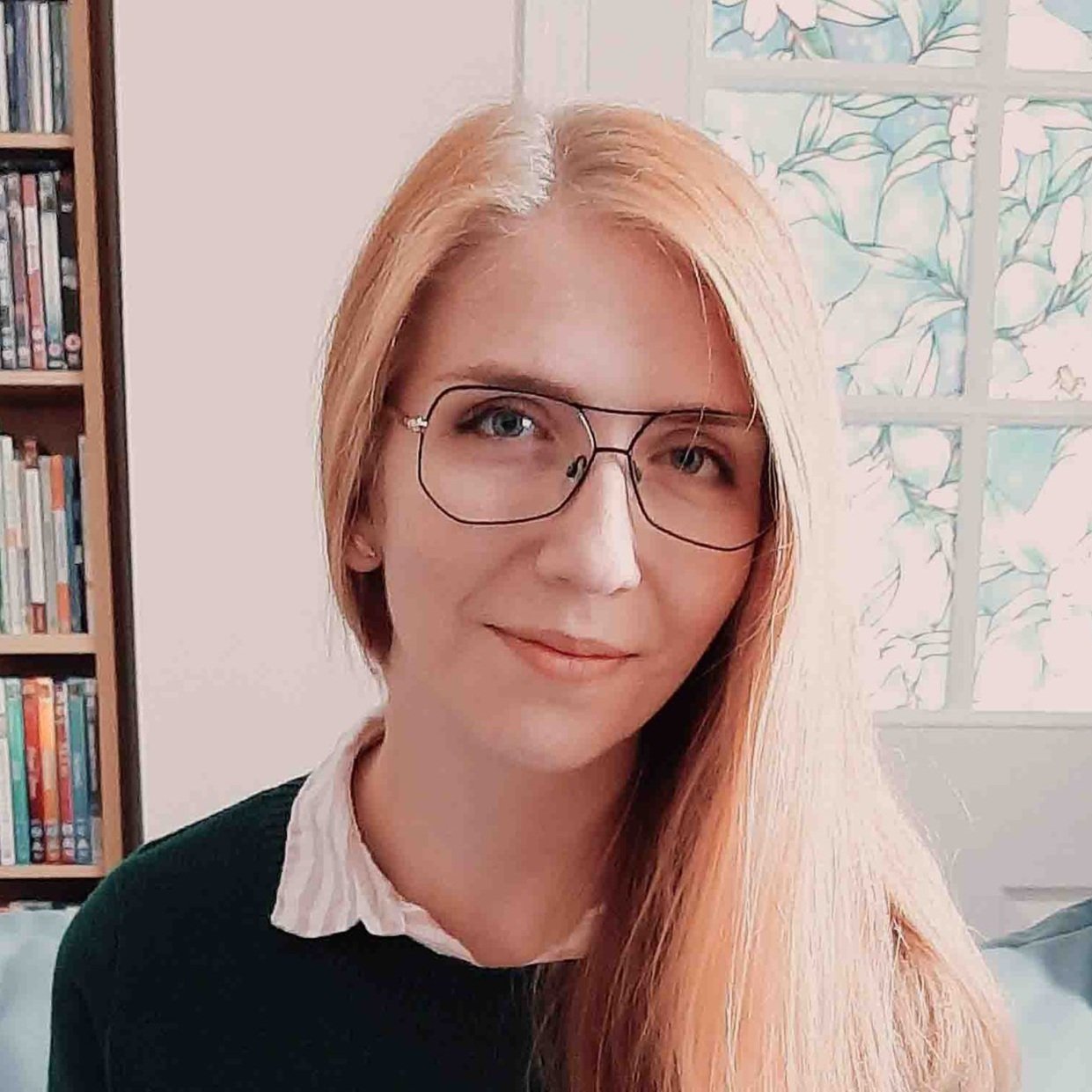 Person with long light hair and glasses, wearing a collared shirt and dark sweater, with a bookshelf and stained glass window in the background.