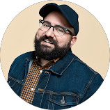 A man with glasses and a beard wearing a baseball cap, denim jacket, and checkered shirt, smiling at the camera