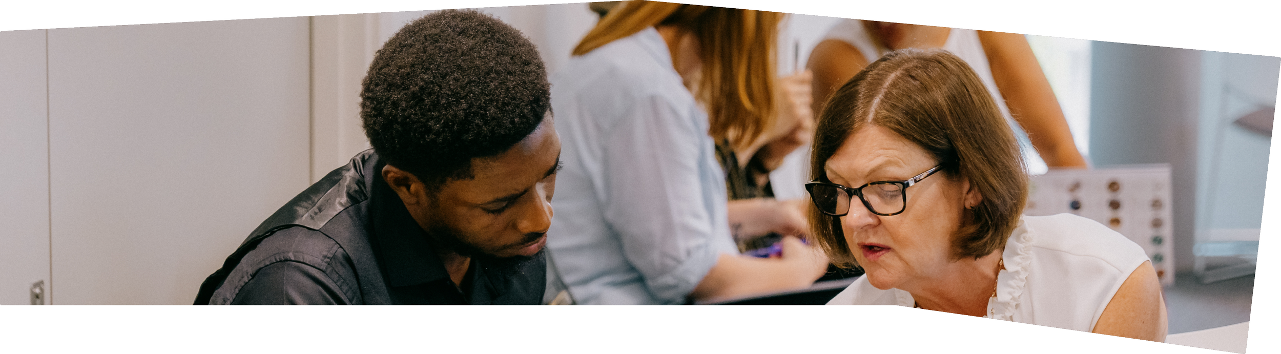 A woman with glasses and a white top is speaking to a young man with dark hair and a dark shirt, in a classroom or office setting, with others working in the background.