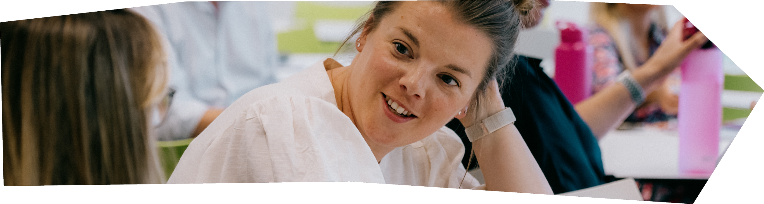 A woman with light skin, brown hair tied up, smiling, looking at a person with long brown hair, who is slightly blurred. The setting appears to be a classroom or study group.