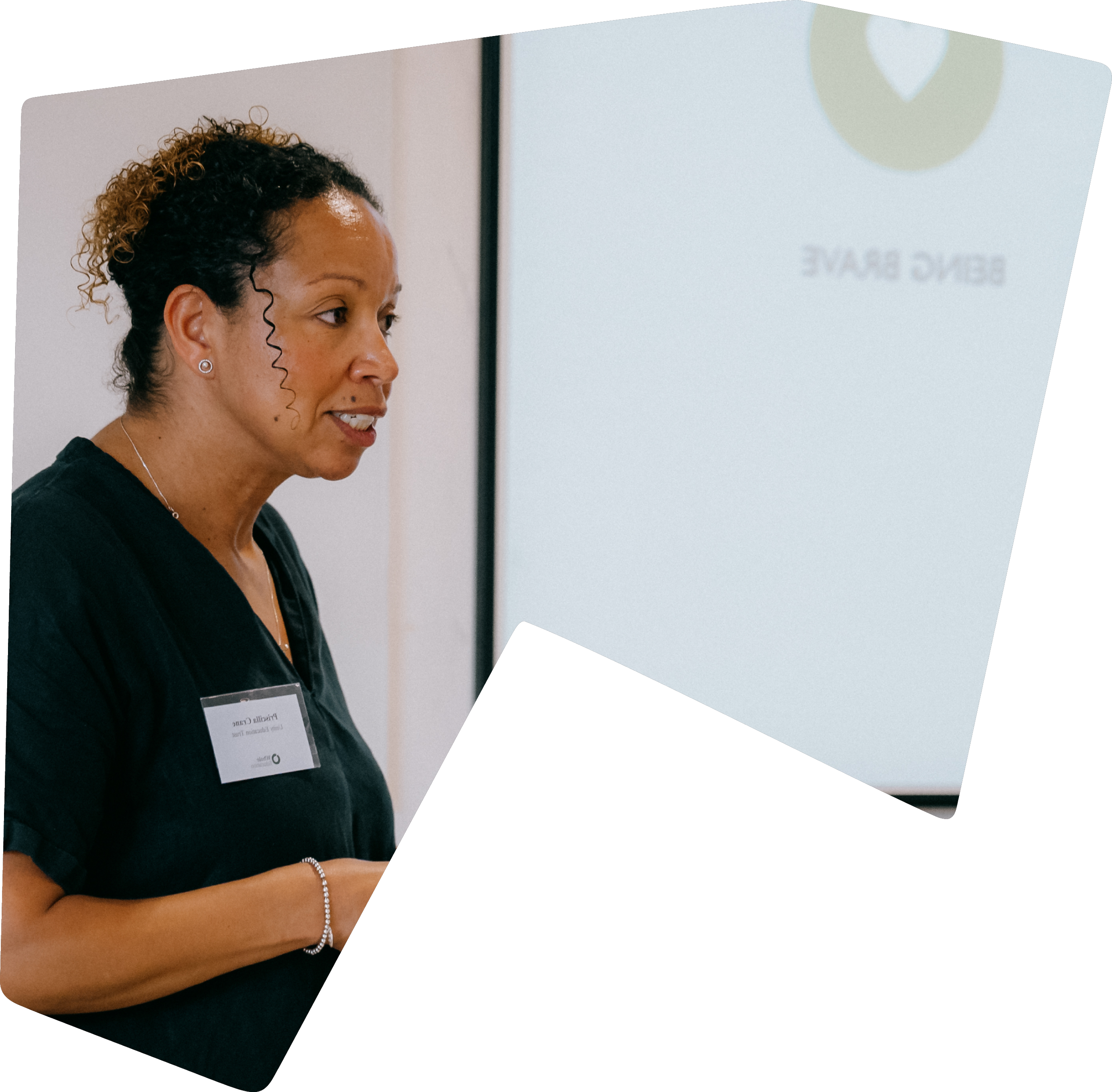 A woman with curly hair and wearing a name tag is speaking or presenting, standing in front of a screen displaying the text 'BING BAVE'.