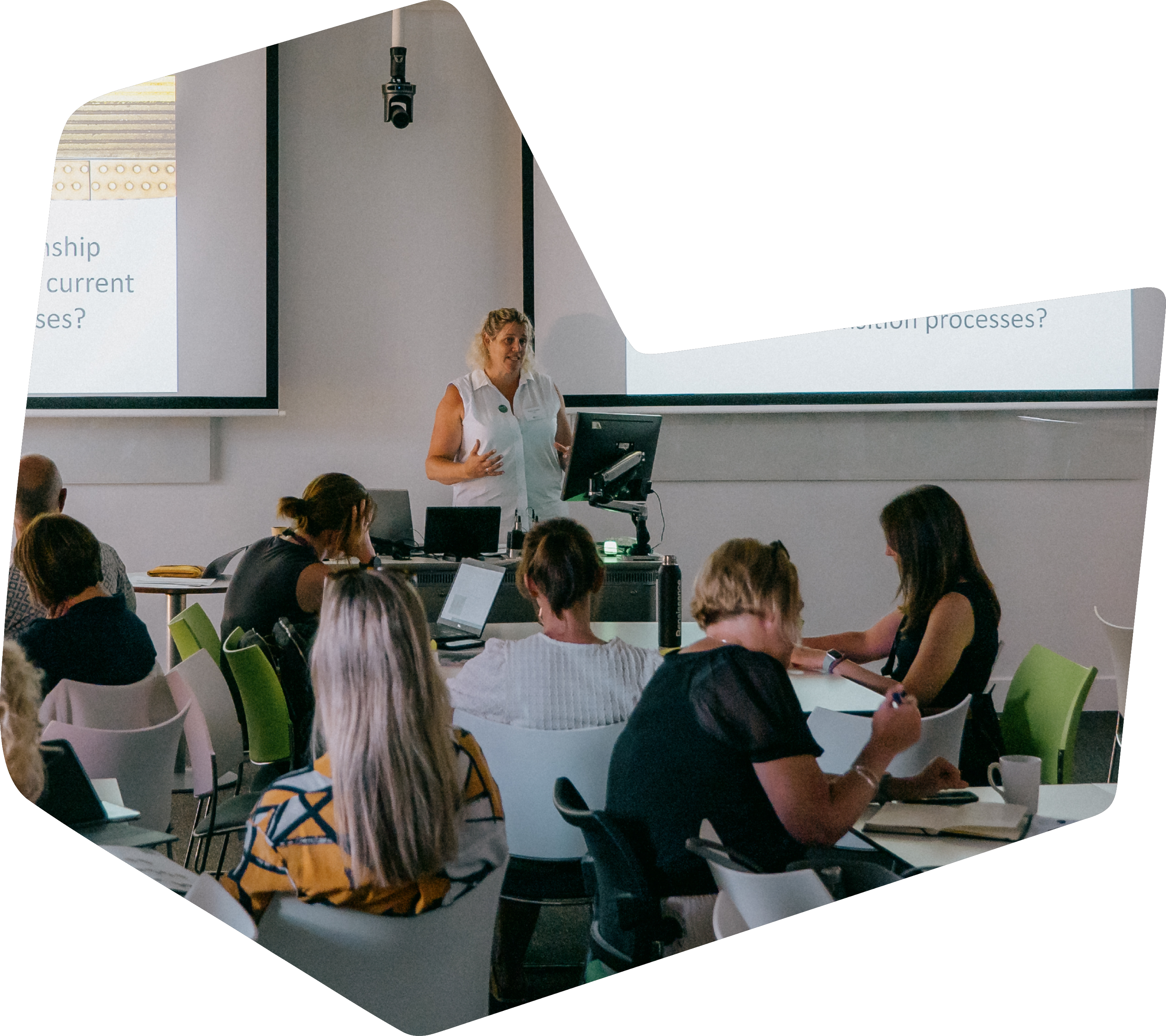 A woman giving a presentation in a classroom with students sitting at tables working on laptops and taking notes.