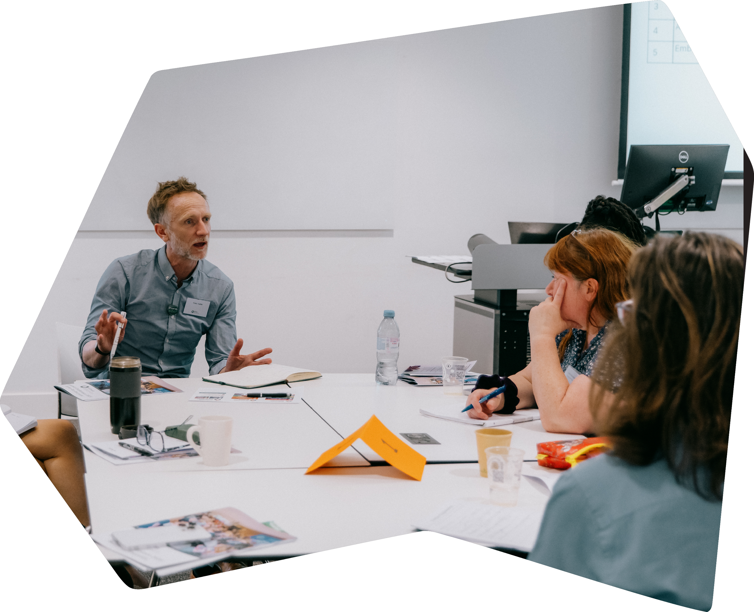 A man in a gray shirt speaking to a group of women in a meeting room.