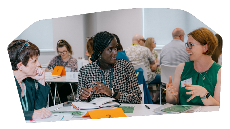 Three women engaged in conversation at a professional conference or workshop with multiple attendees seated at tables in the background.