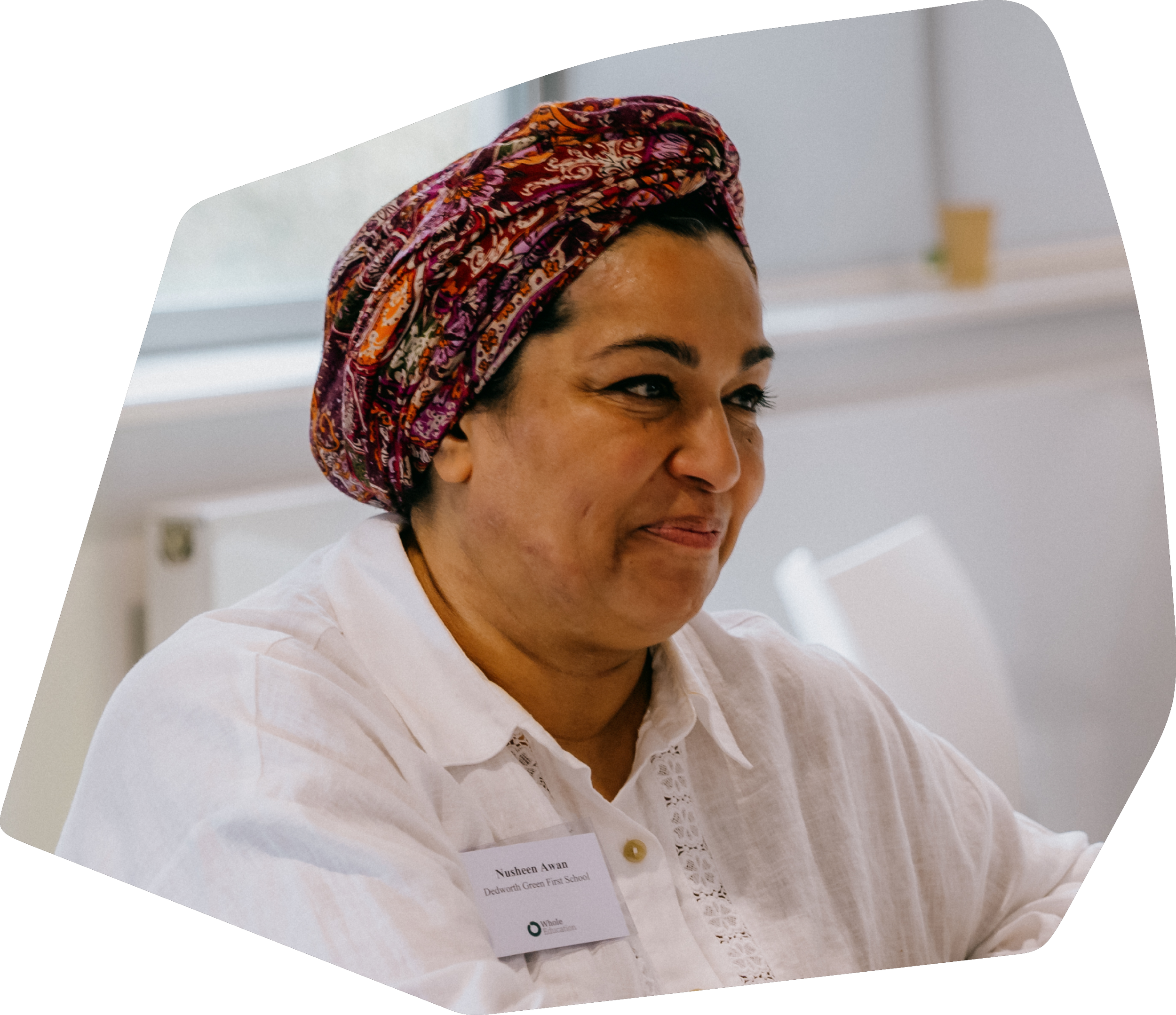 A woman with a colorful headwrap, wearing a white shirt with a name tag, sitting indoors and smiling.