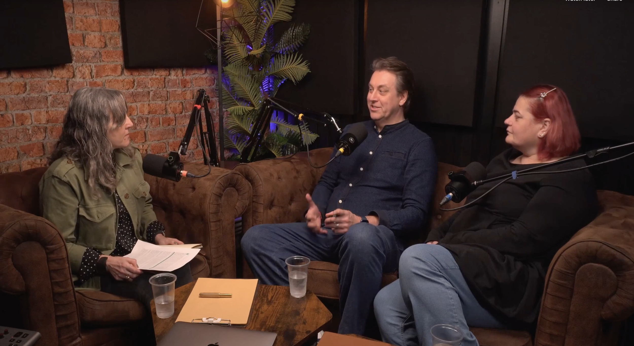 Three people having a conversation in a podcast studio with microphones, sitting on leather couches, brick wall background, and plants.