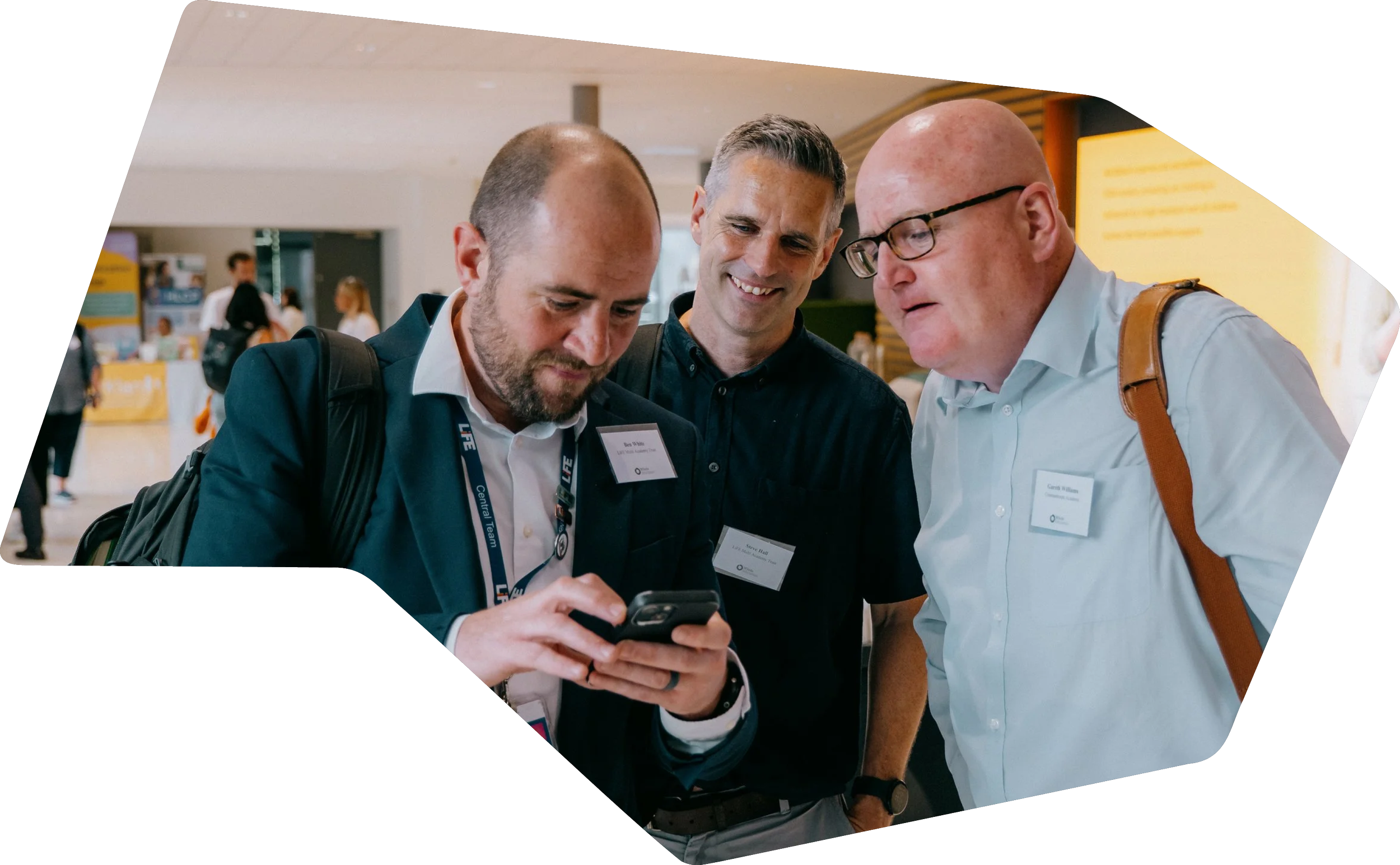 Three men examining a smartphone together at a conference, with conference badges and lanyards, in a busy indoor setting.