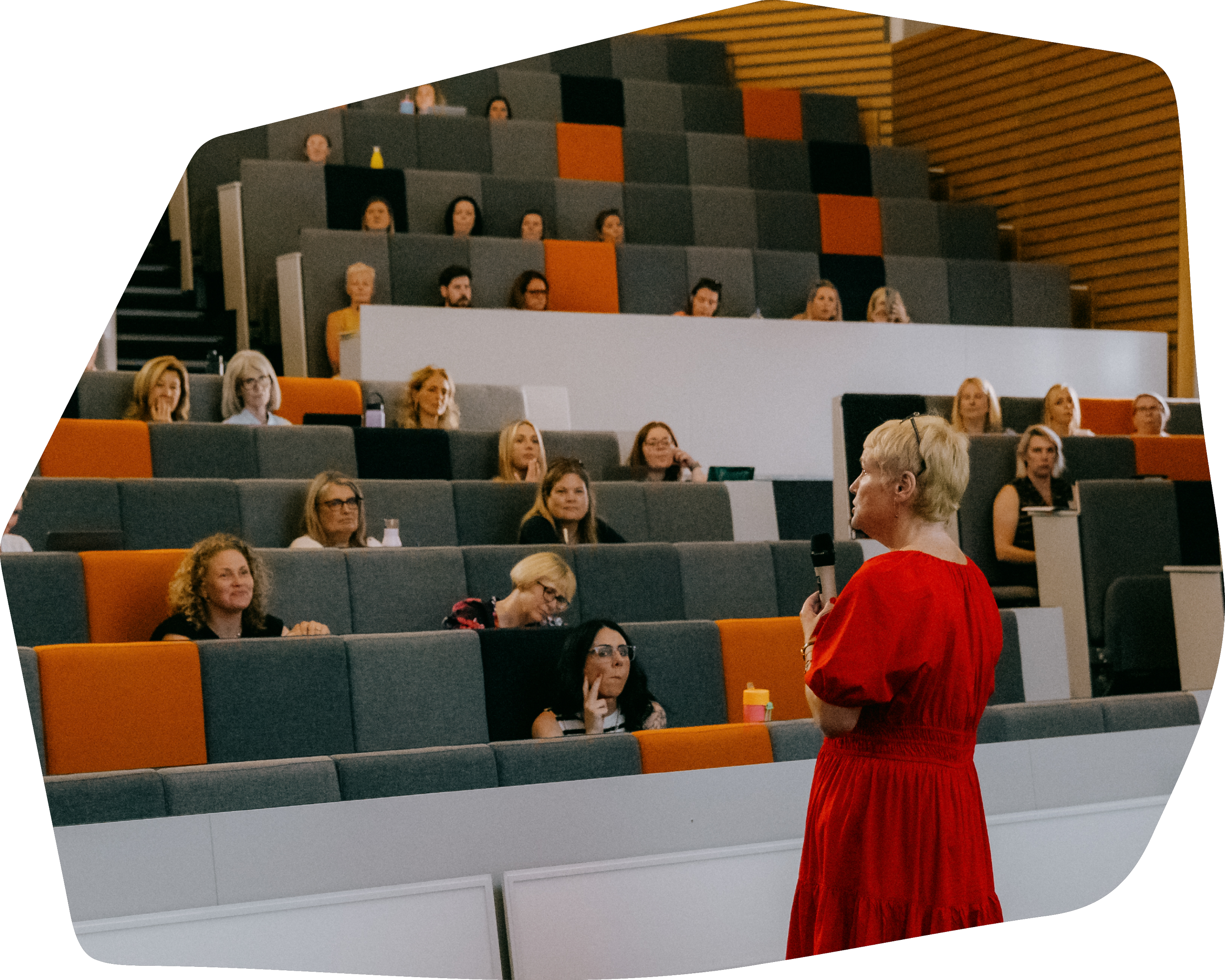 A woman in a red dress giving a presentation or speech in front of an audience in a modern lecture hall. The audience consists of women seated in tiered rows, attentively listening.