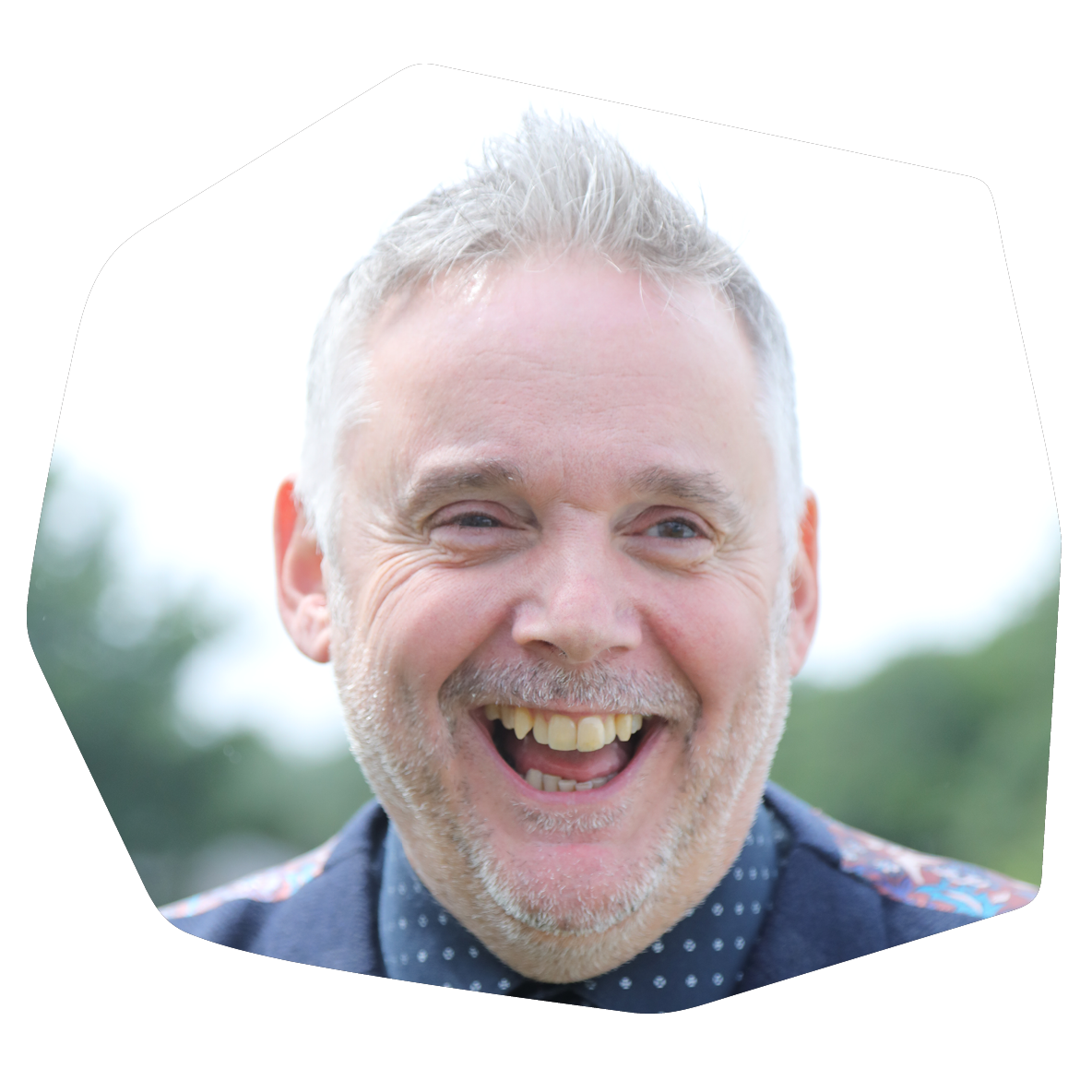 A middle-aged man with gray hair and a beard, smiling widely outdoors with a blurred background of trees and sky.