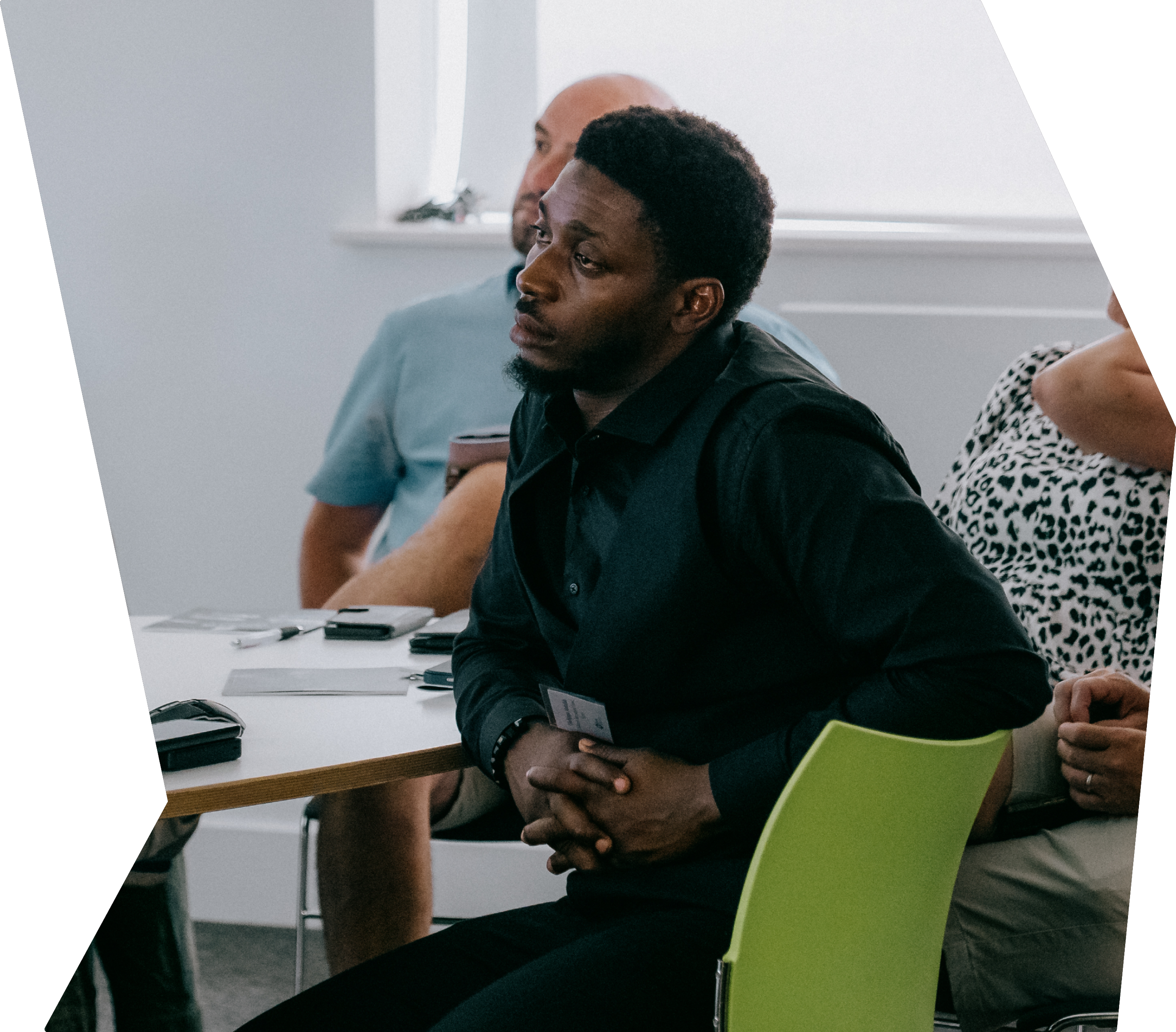 People attending a conference or meeting, sitting at a table with notebooks and electronic devices, listening attentively.