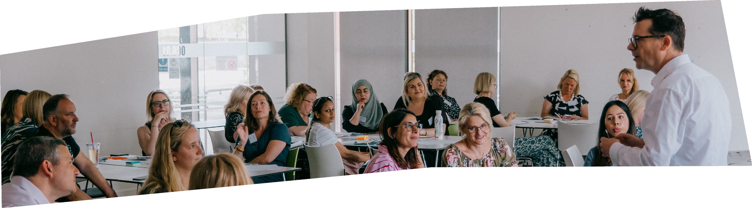 A man in a white shirt and glasses speaking to a large group of women and a few men in a conference room with large windows.