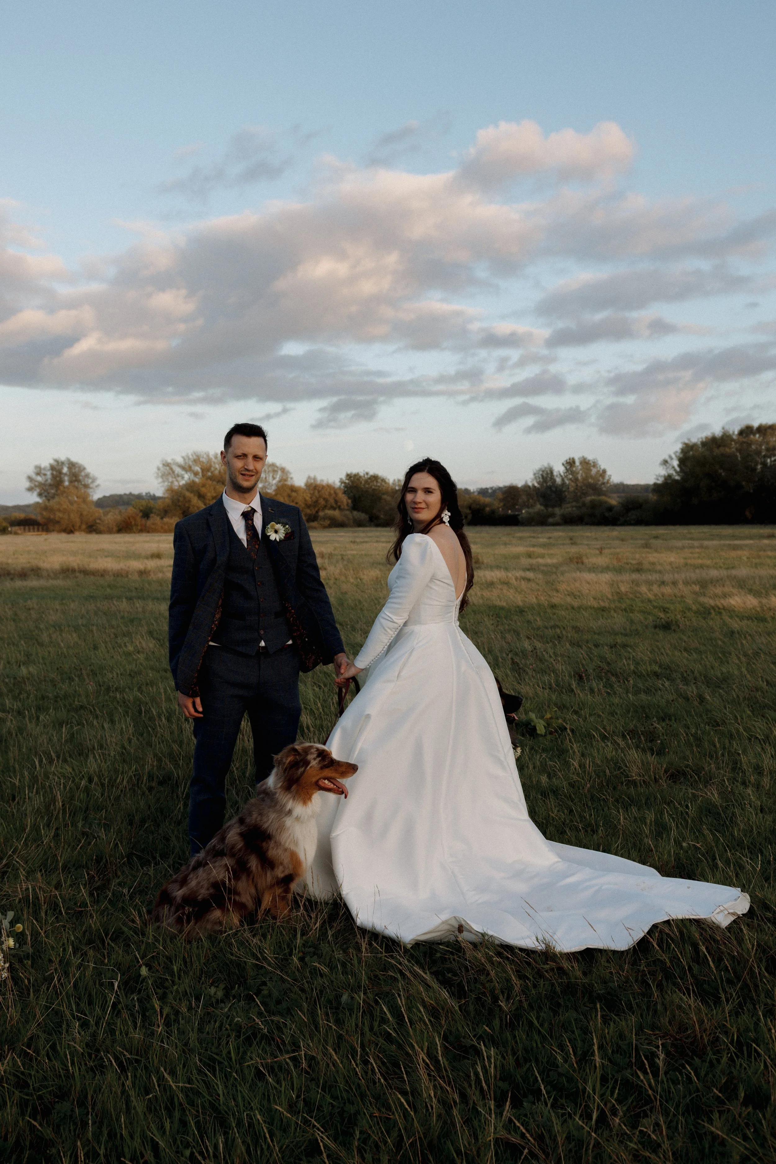 A bride and groom holding hands in a grassy field with a dog, under a cloudy sky, during sunset.