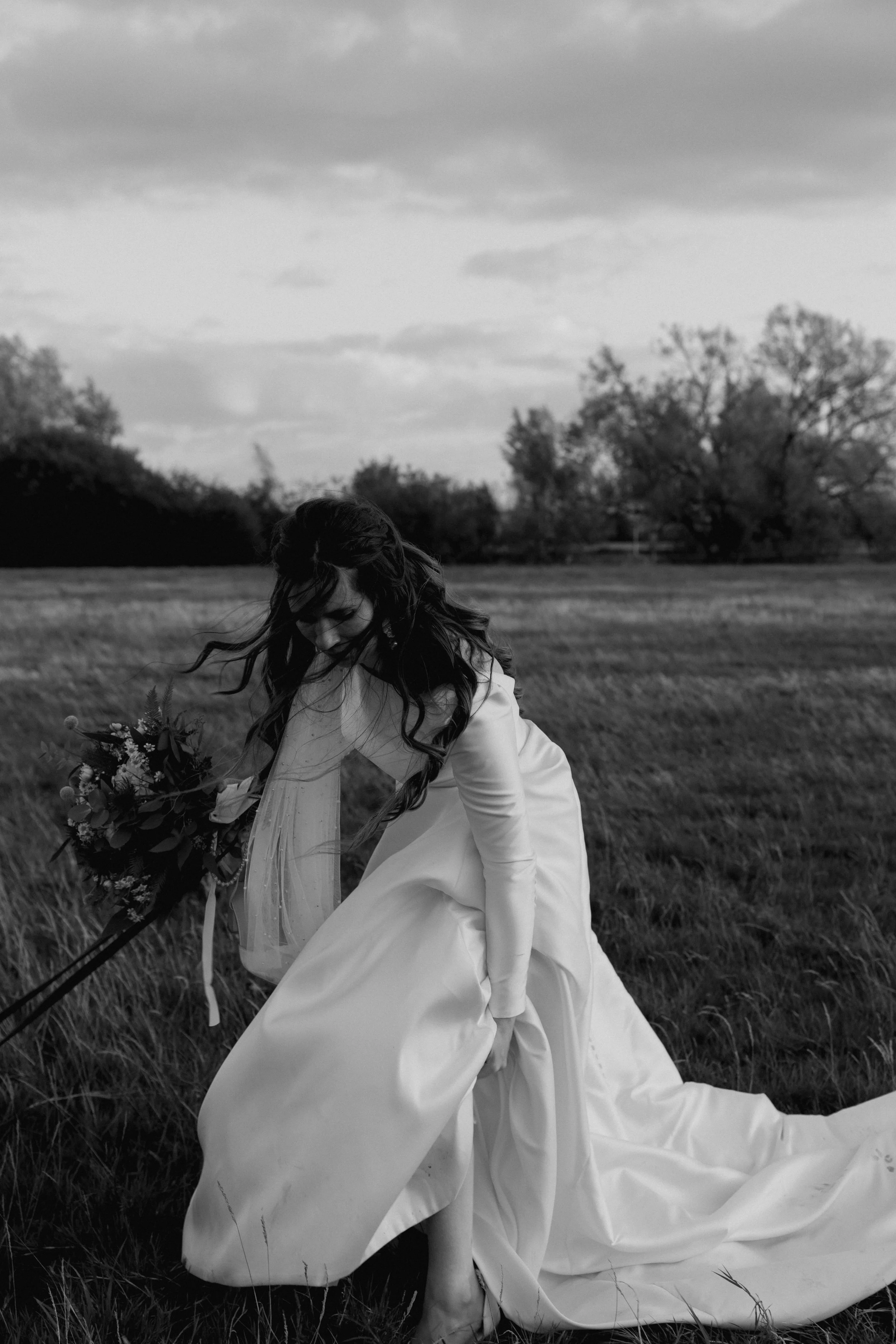 A woman in a wedding dress holding a bouquet, bending down in a grassy field outdoors, with trees and cloudy sky in the background, in black-and-white photograph.