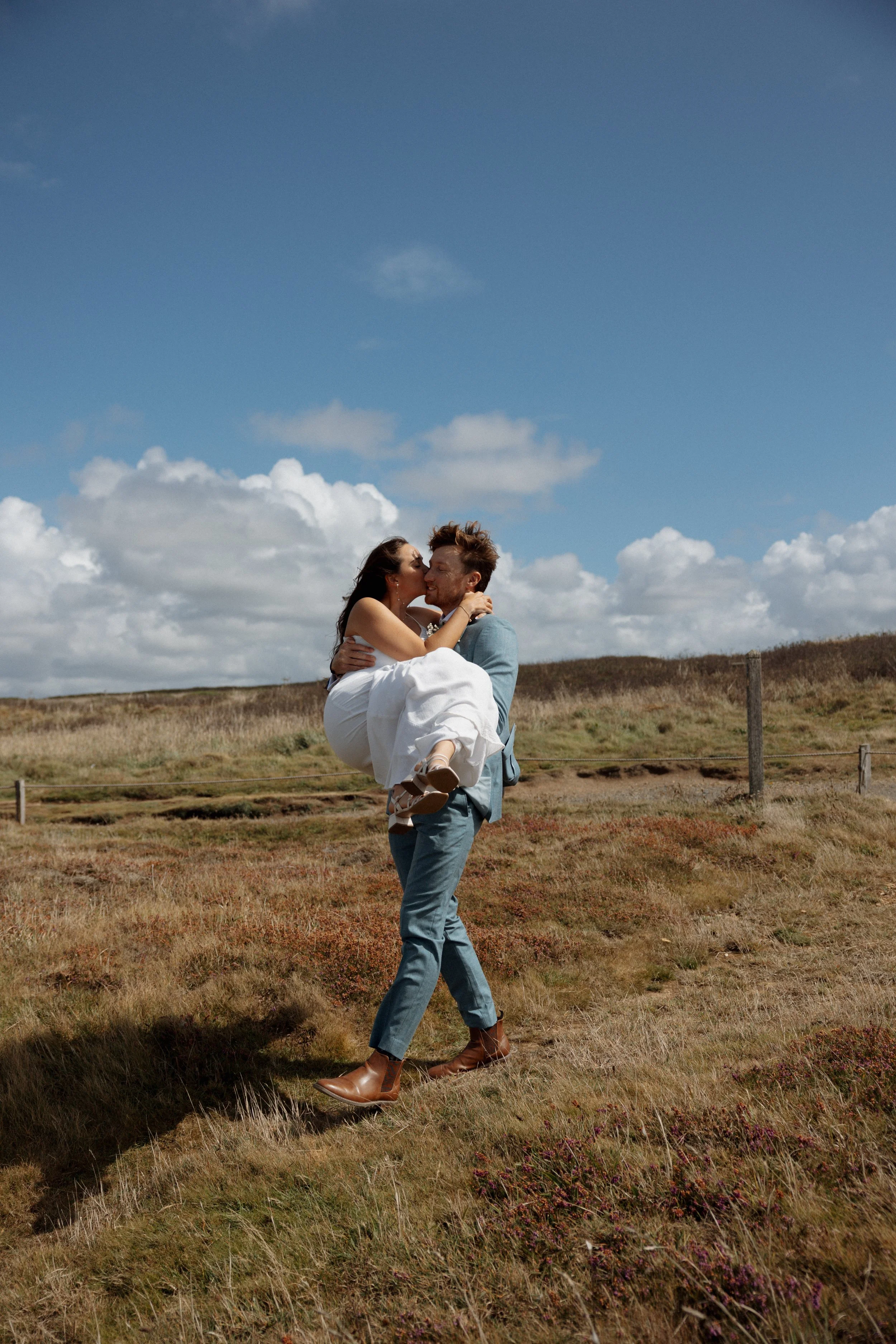 A man carrying a woman in his arms outdoors on a grassy field, under a cloudy blue sky, both smiling and about to kiss.