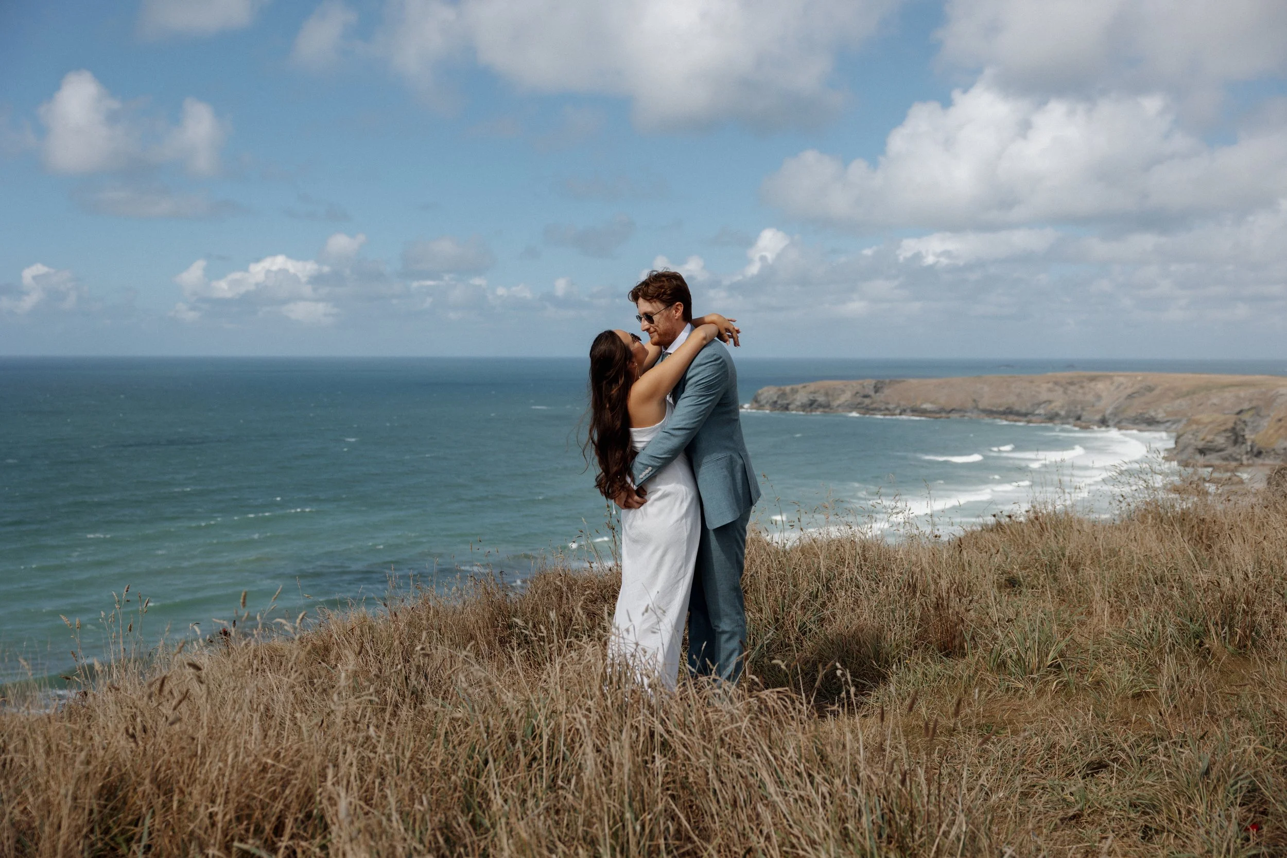 A couple embracing on a grassy cliffside overlooking the ocean with cliffs in the distance under a partly cloudy sky.