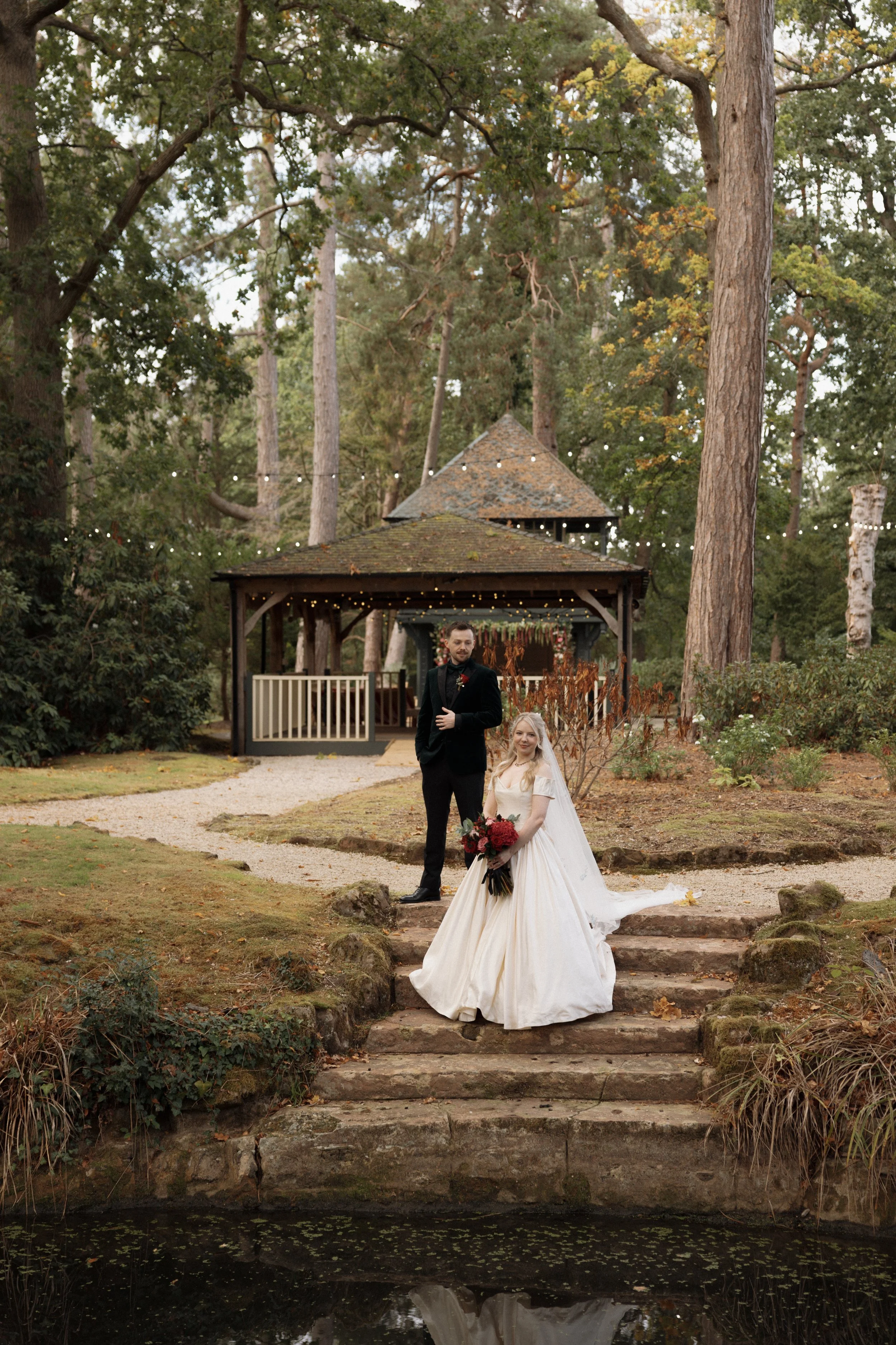 Bride and groom pose beside a rustic pavilion decorated with string lights in a wooded garden.