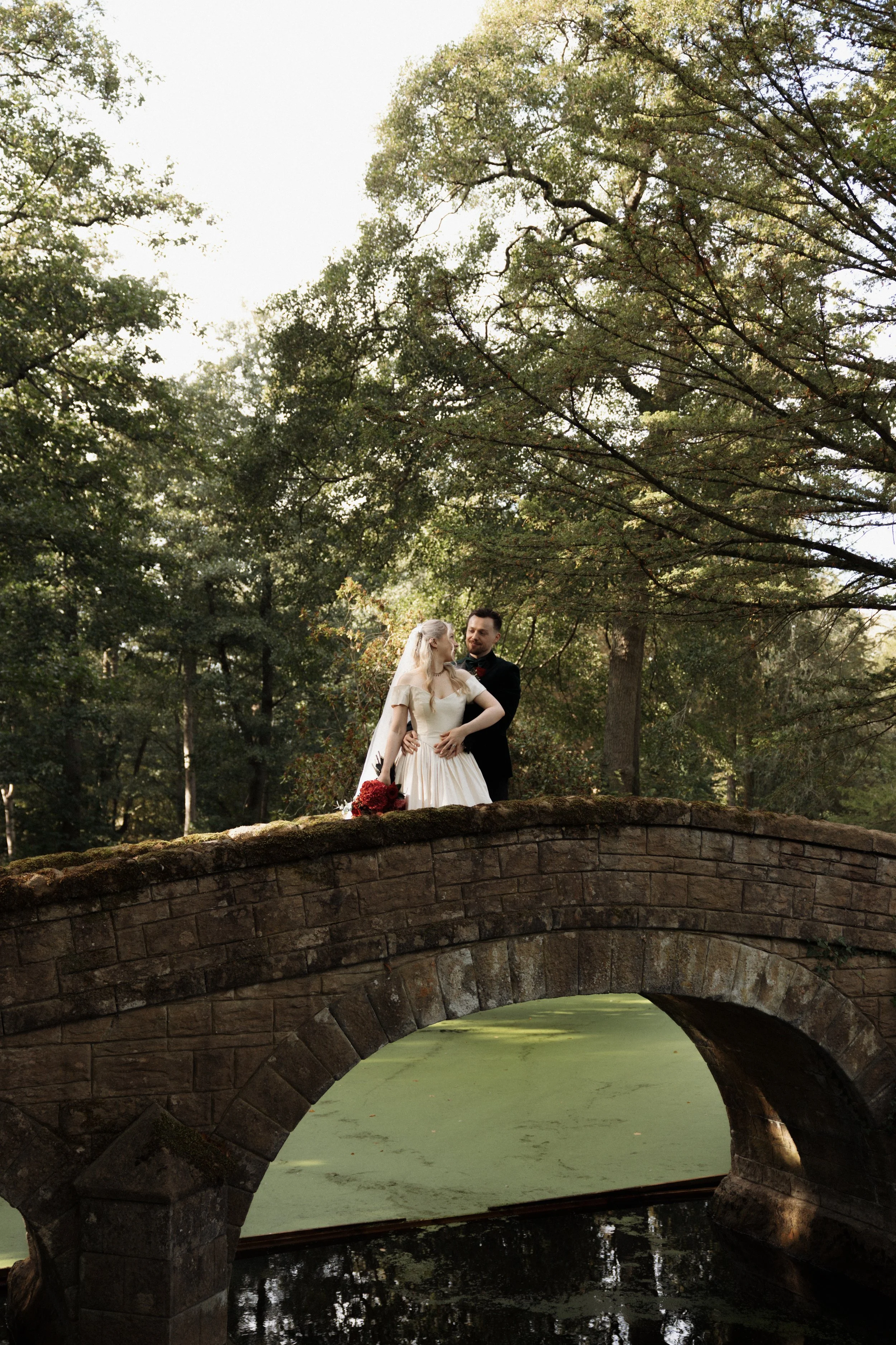 Bride and groom embracing on a moss-covered stone bridge in a wooded park, with the bride holding a red flower bouquet.