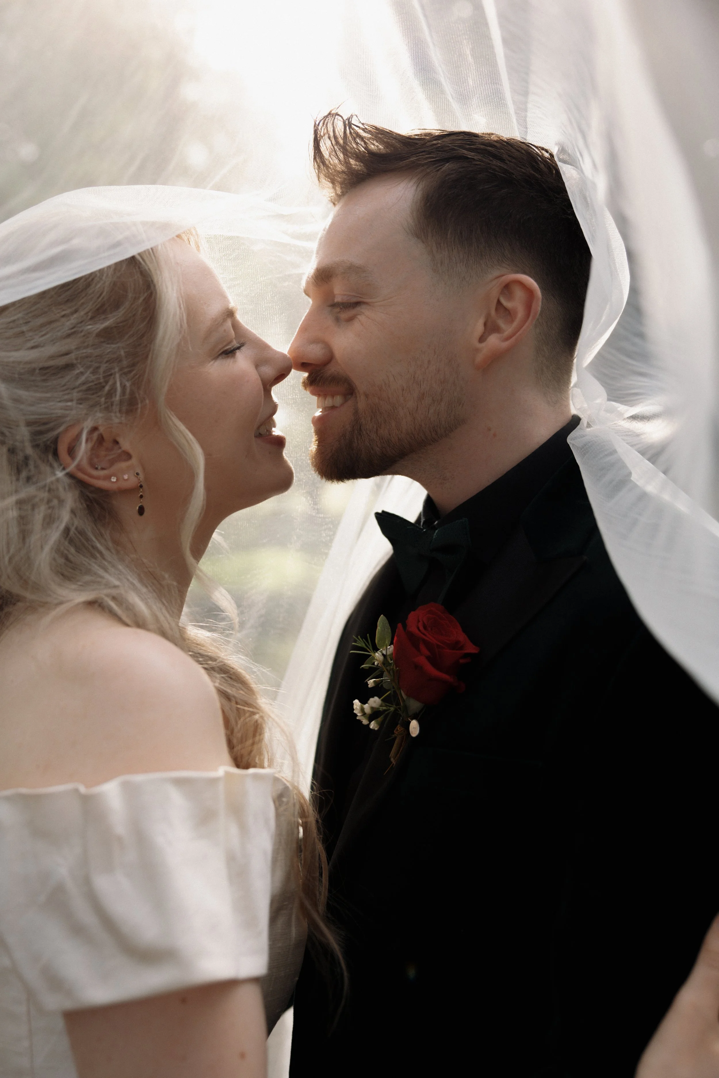 Newlywed couple smiling and touching noses beneath a white veil, with the groom wearing a red rose boutonniere.
