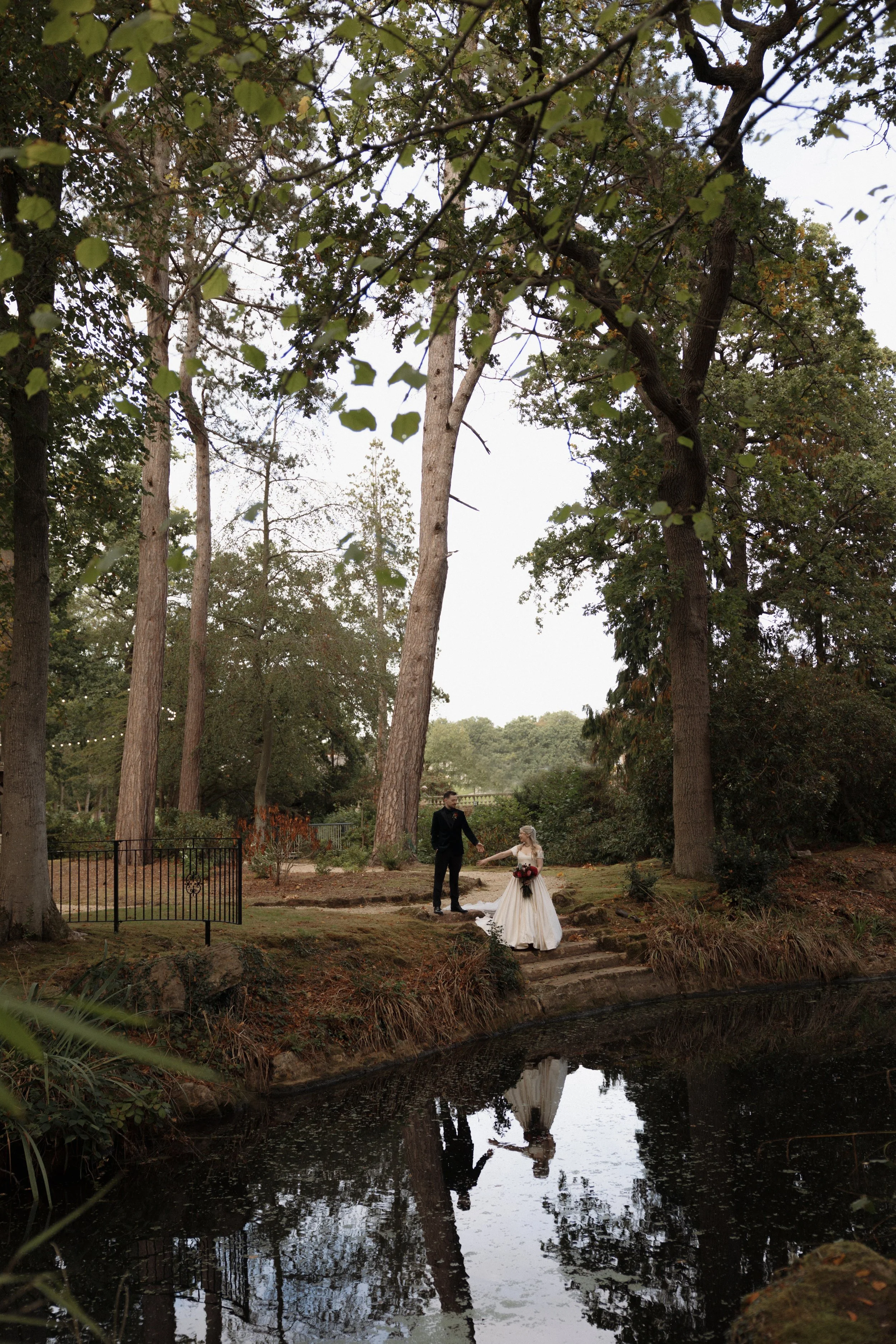 Bride and groom posing beside a pond in a wooded garden, surrounded by tall trees and reflected in the water.