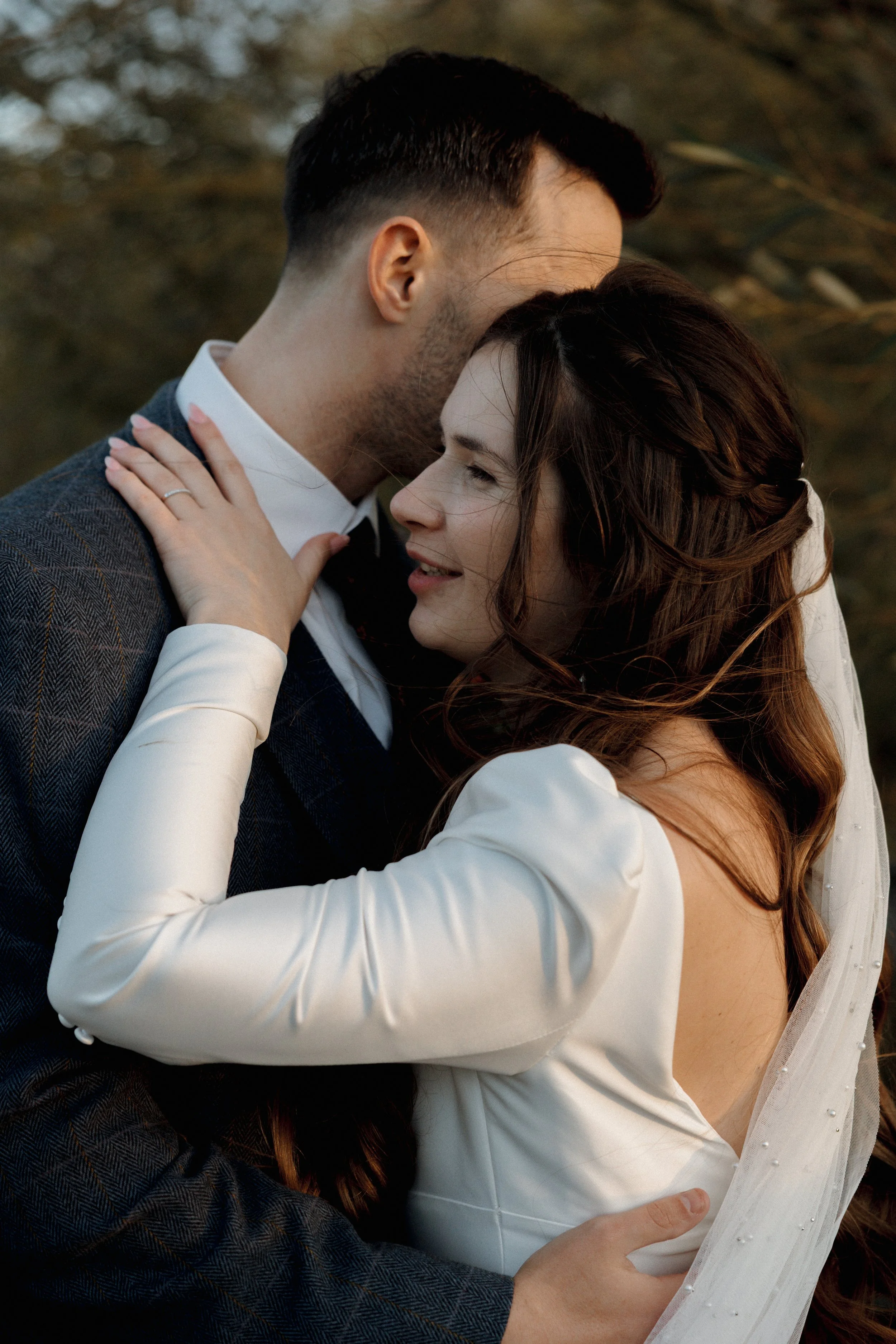 A bride and groom embracing closely outdoors during their wedding, with the bride wearing a white dress and veil, and the groom in a dark suit.