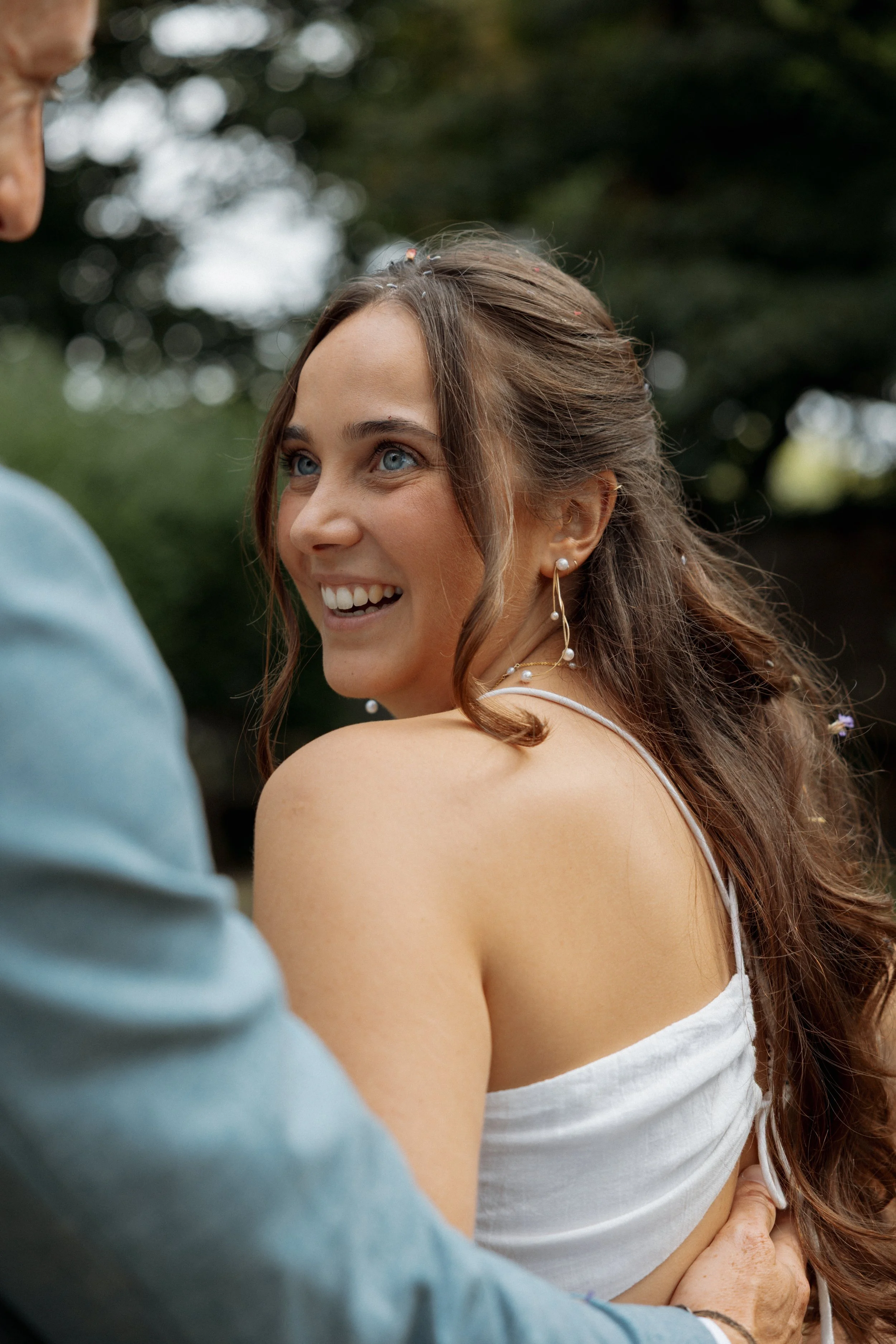 A woman with long, wavy brown hair and blue eyes smiling as she looks over her shoulder, wearing pearl earrings and a white top, outdoors with a blurred green background.