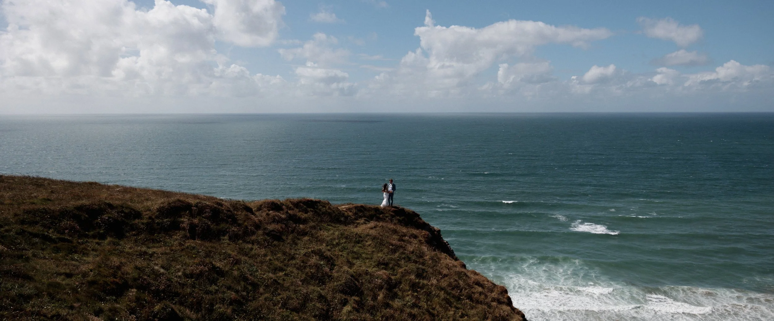 A couple stands on a cliff overlooking the ocean, under a partly cloudy sky.