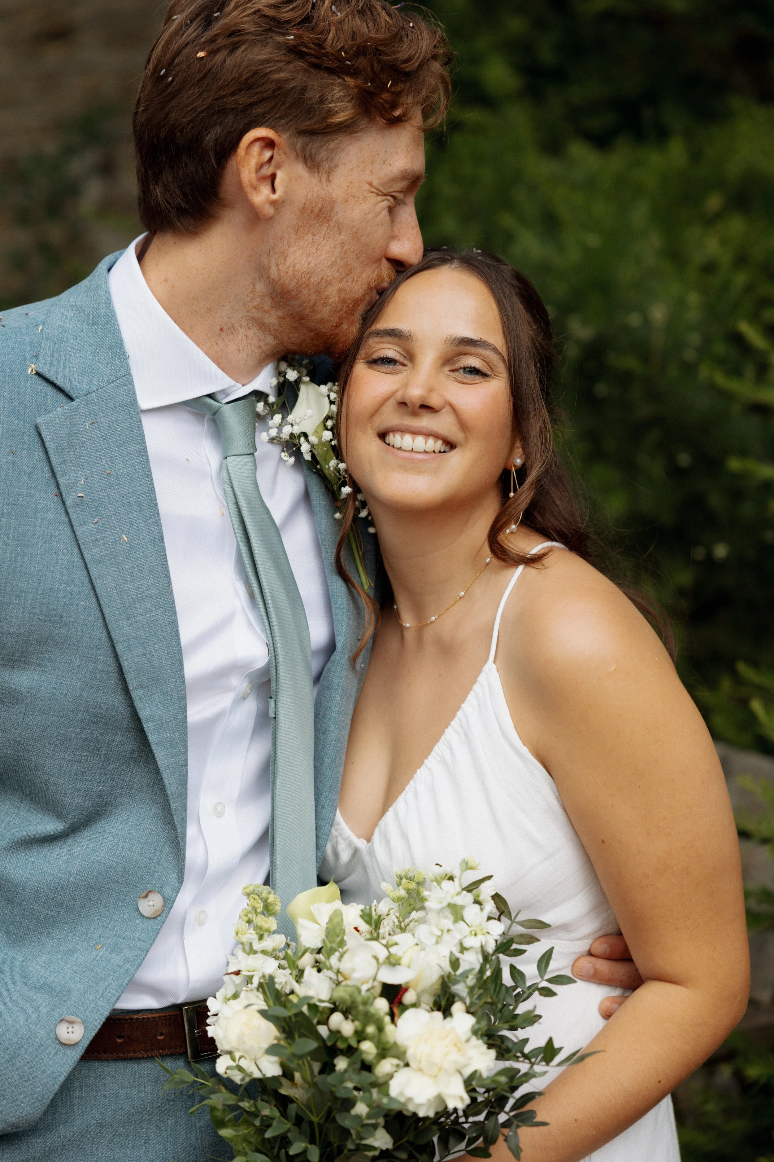 A newlywed couple outdoors, with the groom kissing the bride on the forehead. The bride is smiling while holding a bouquet of white flowers, wearing a white dress, and the groom is in a light blue suit with a tie.
