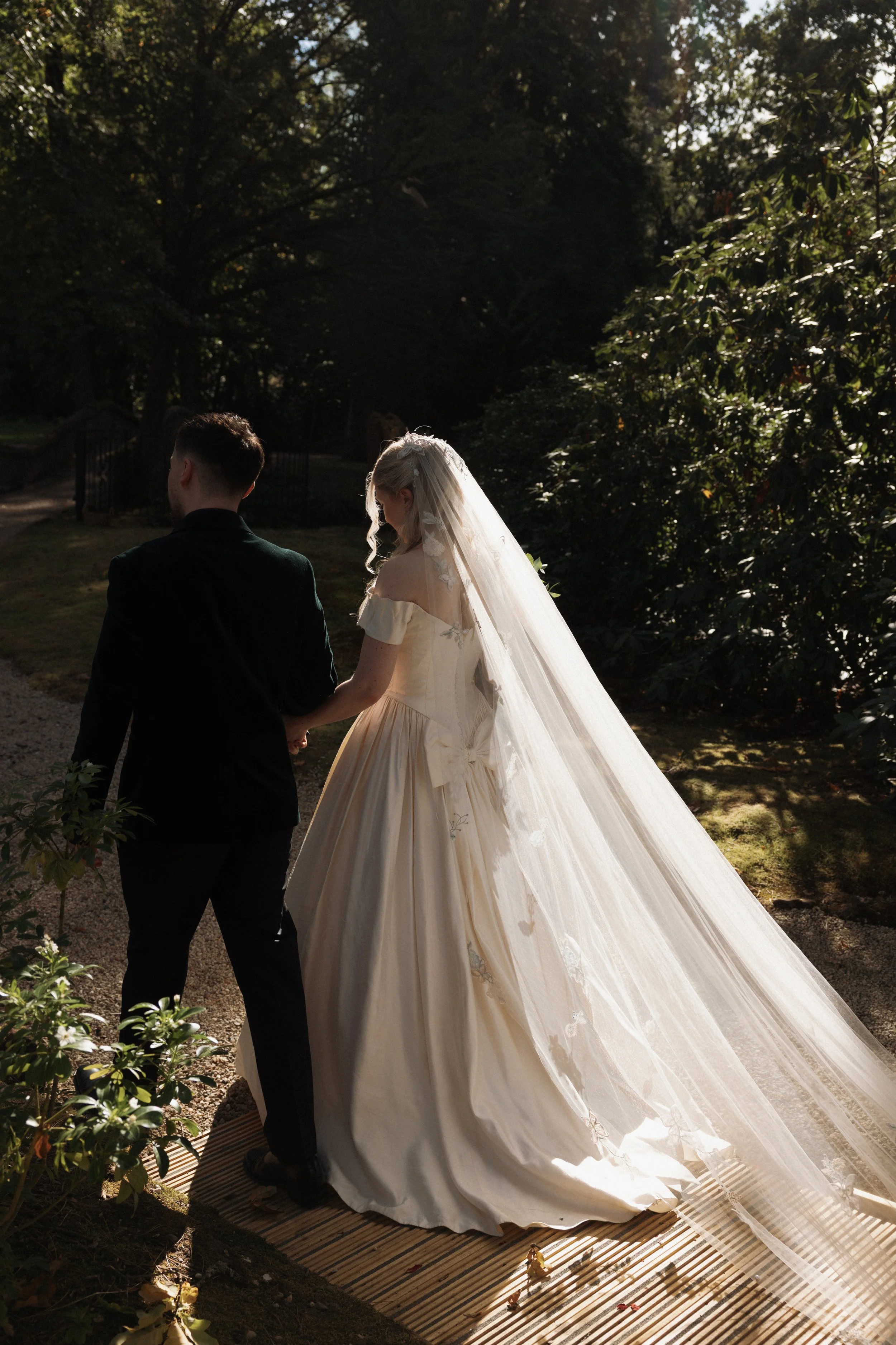 Newlywed couple walking hand in hand through a sunlit garden, with the bride’s long veil trailing behind her.