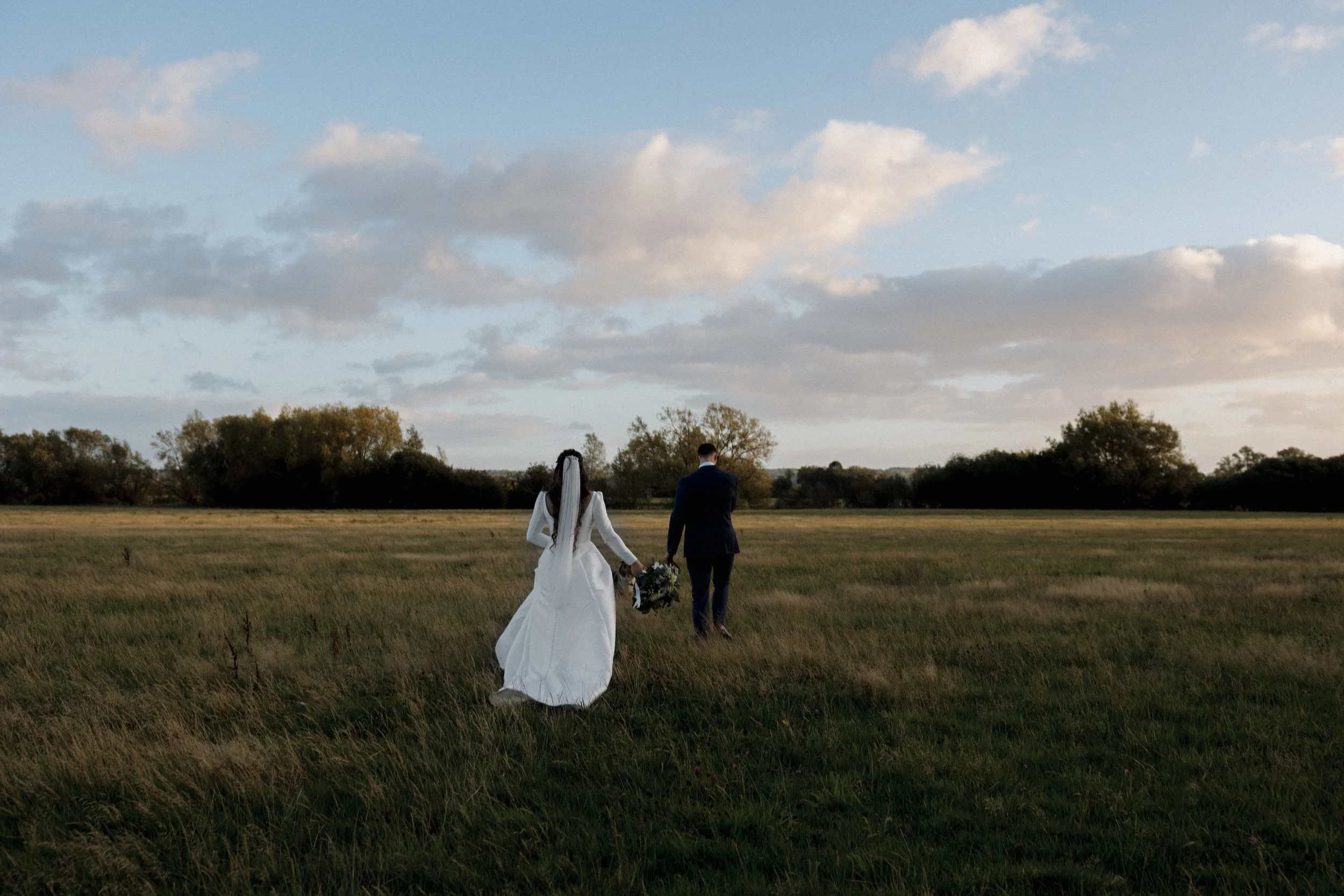 A couple walking through a grassy field, holding hands, during sunset with clouds in the sky.