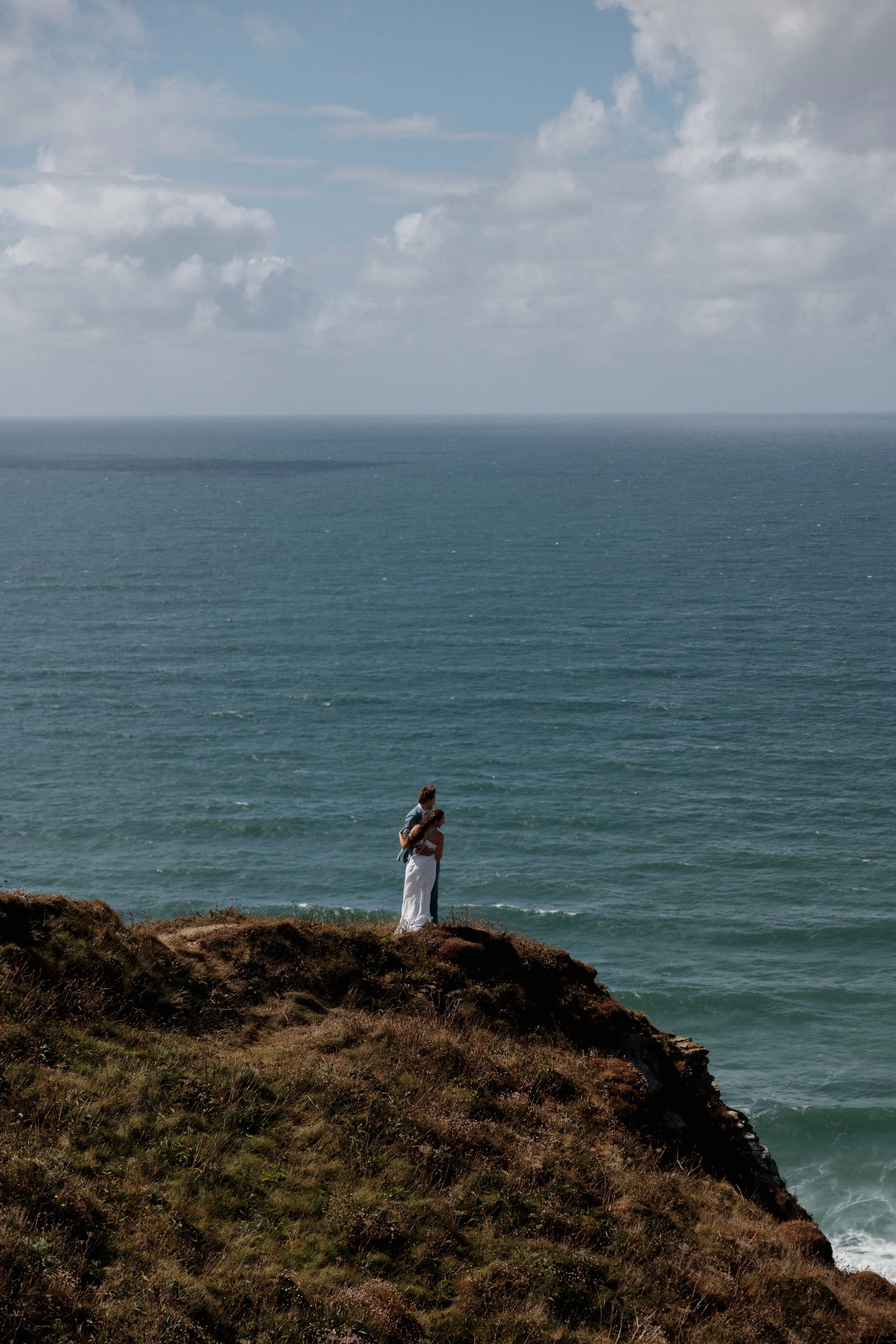 A couple standing on a grassy cliff overlooking the ocean, with a cloudy sky above.
