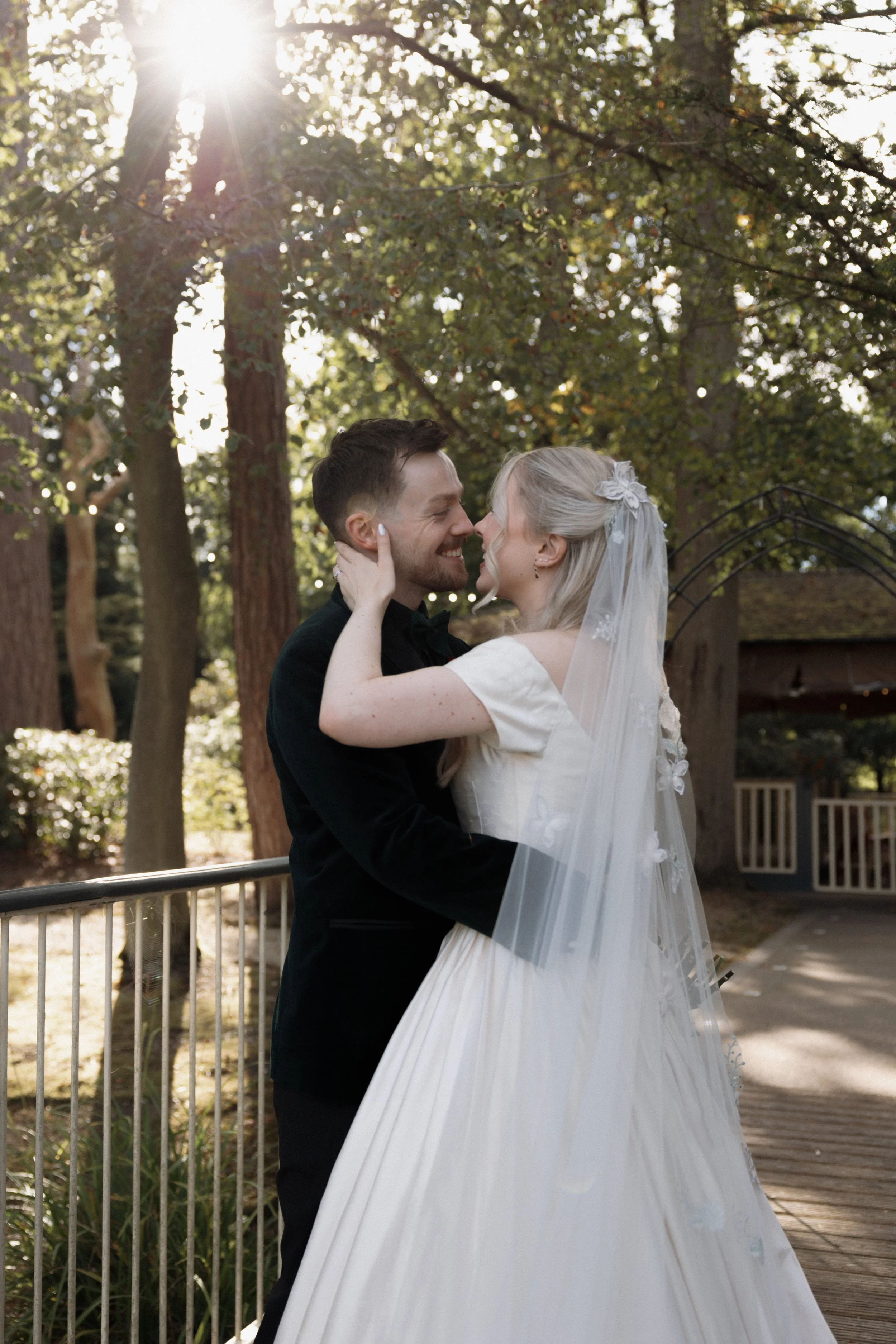 Newlywed couple kissing outdoors on a sunlit forest deck, with the bride in a white wedding dress and veil.