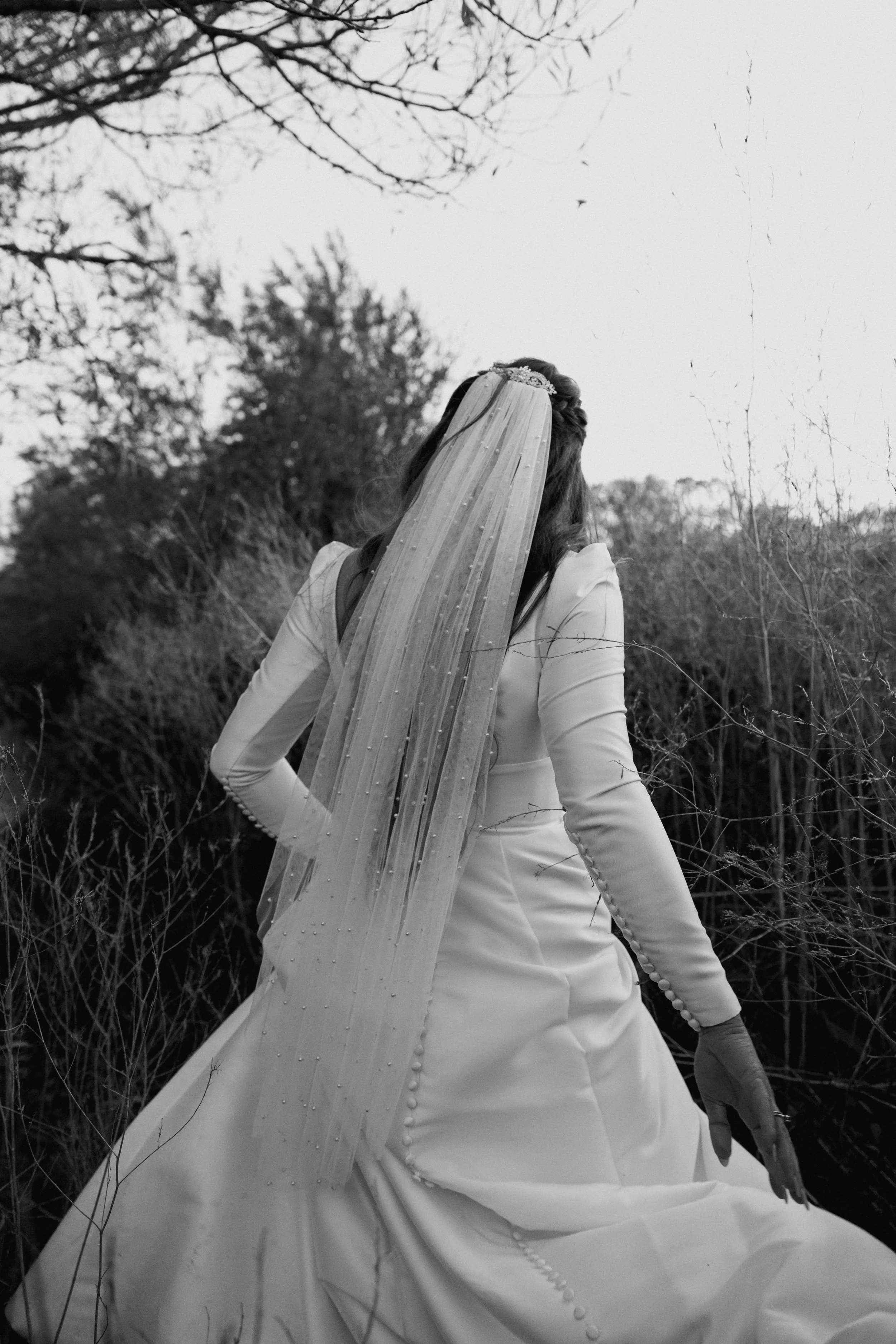 A woman in a wedding dress with a veil outdoors, surrounded by branches and trees, in black and white.