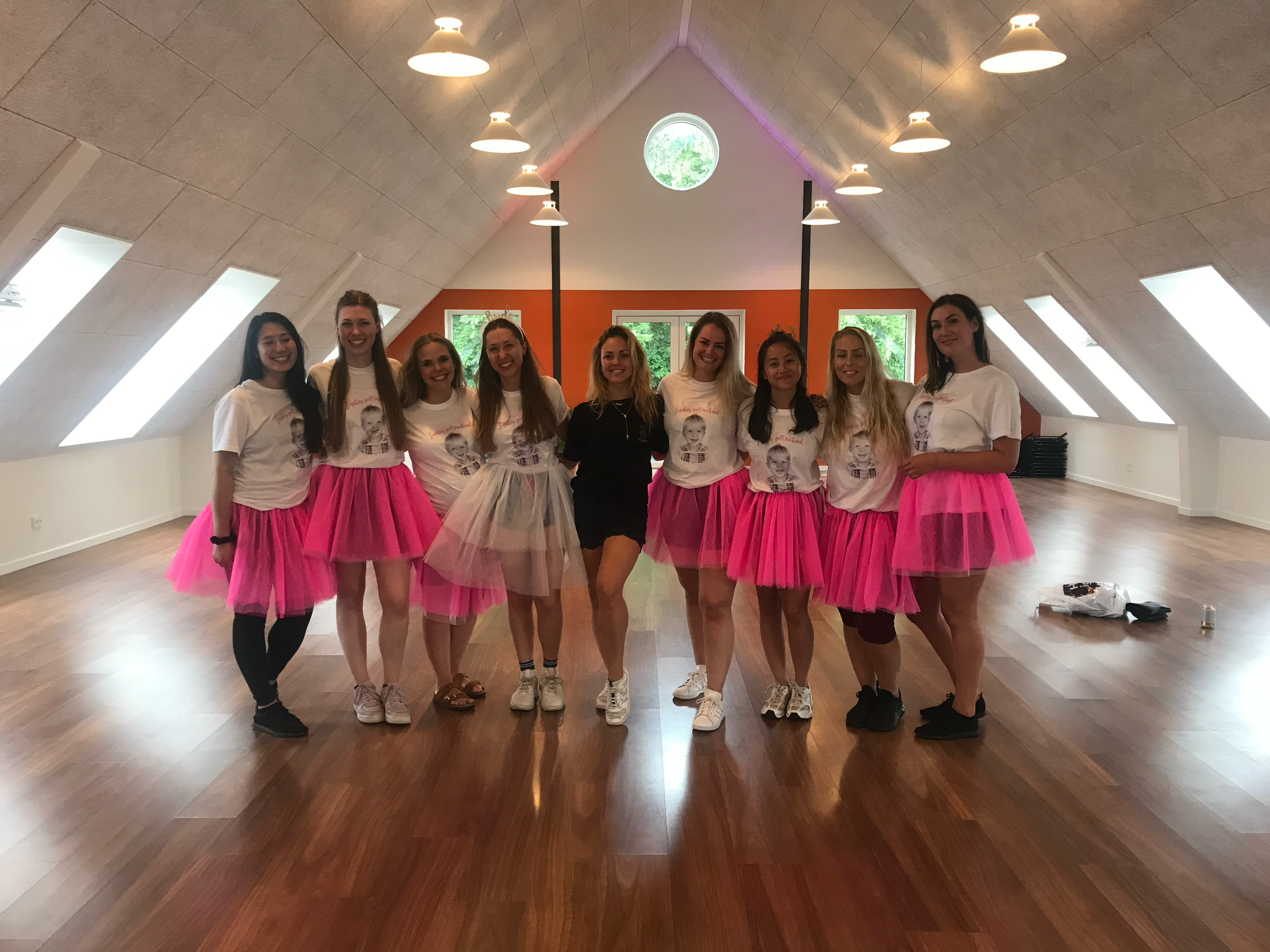 En gruppe kvinder, nogle i hvide T-shirts med billeder og teksten 'Proud to be a woman', og andre i pink kjoler, poserer for en gruppefotos i en indendørs lokalitet med træ- og hvide vægge. Alle smiler. Polterabend 