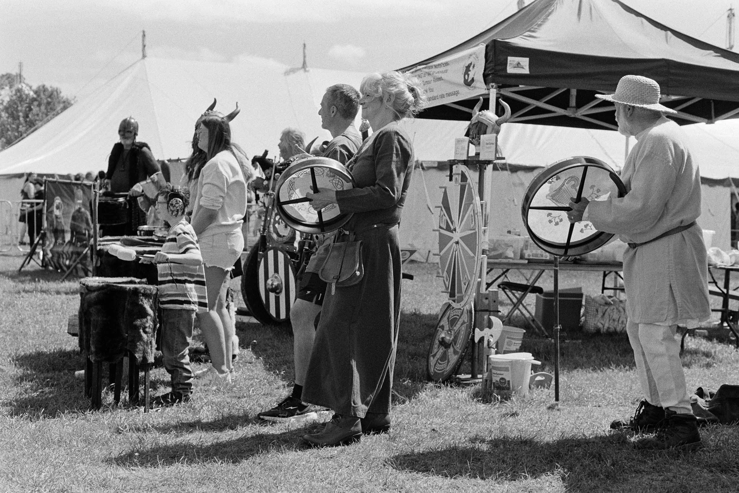 A line of people at an outdoor event, many wearing Viking helmets with horns, some with musical drums, standing under tents with various displays.