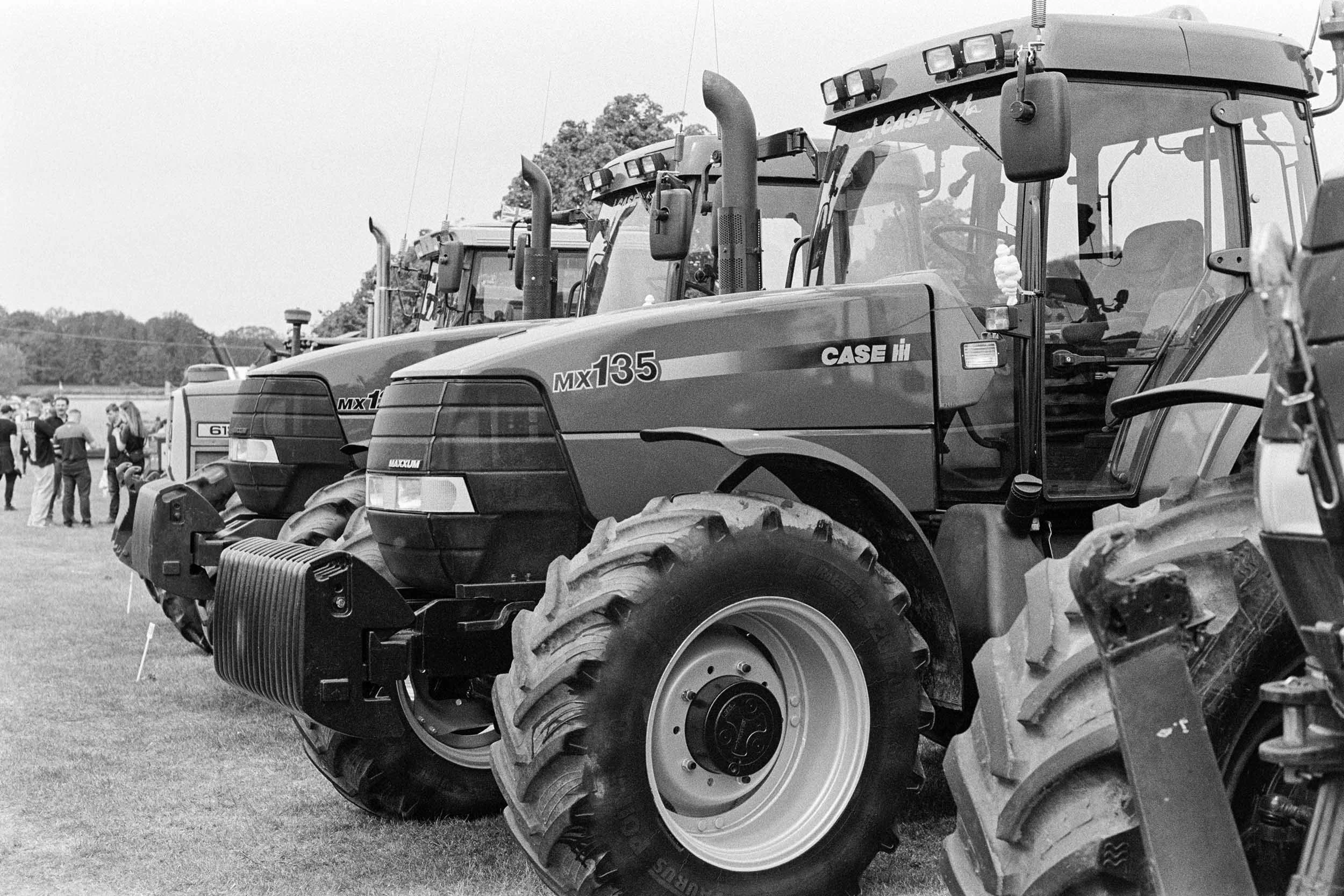 A row of large tractors at an outdoor exhibition, with people walking and observing in the background.