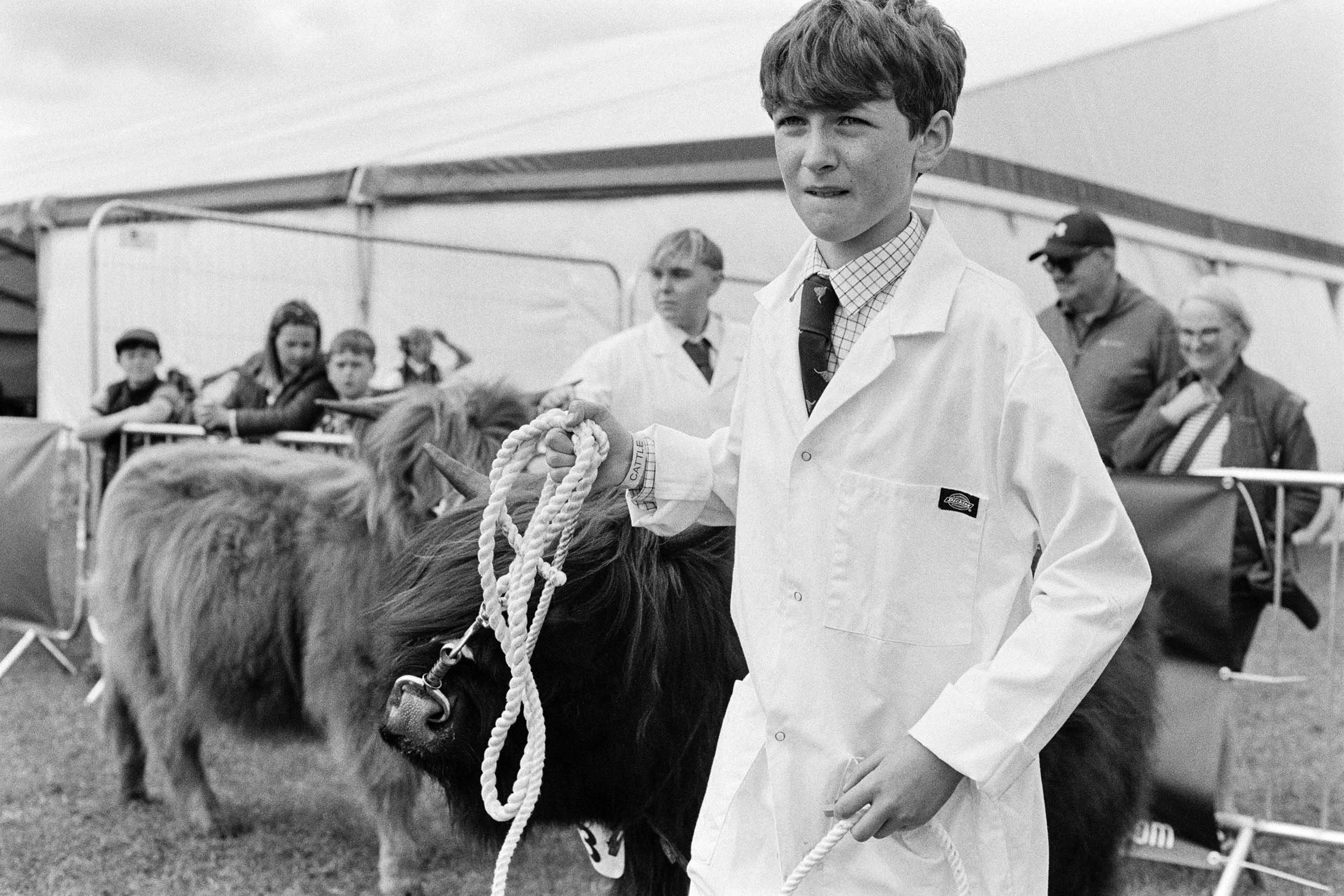 A young boy in a white coat holding a rope attached to a horse at an outdoor event, with several children and adults in the background.
