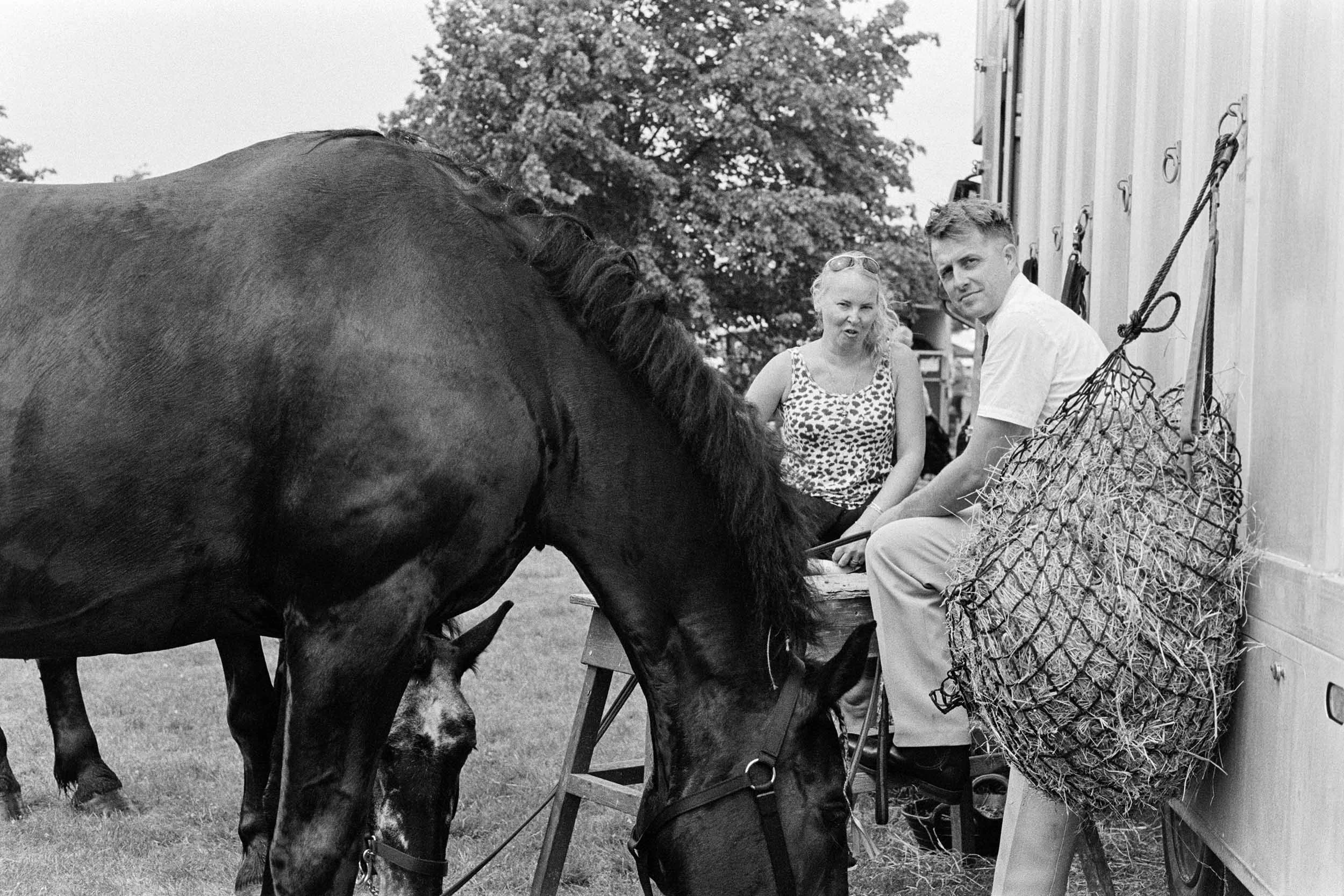 A black and white photo of a woman and a man sitting on folding chairs outside, next to a horse, with trees in the background. The woman is wearing sunglasses on her head and a leopard print sleeveless top, while the man is in a white shirt. There is