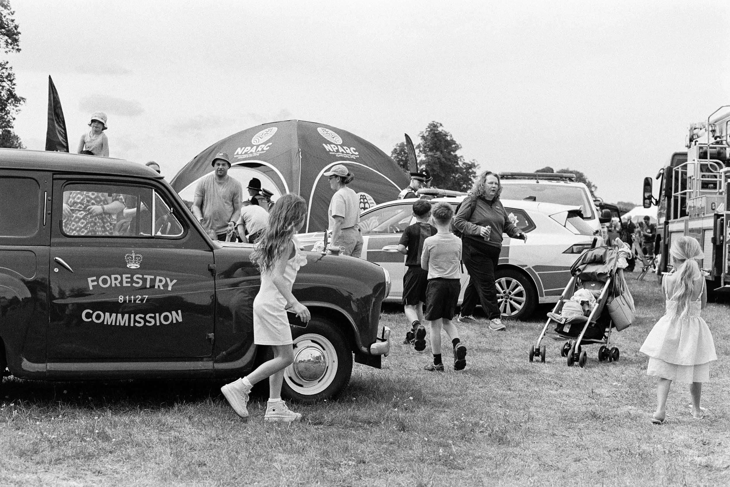 A black-and-white photo of a crowd at an outdoor event with police cars and a tent in the background. People of various ages, including children and adults, are walking around, some talking or holding items. The police vehicle has the words 'Forestry