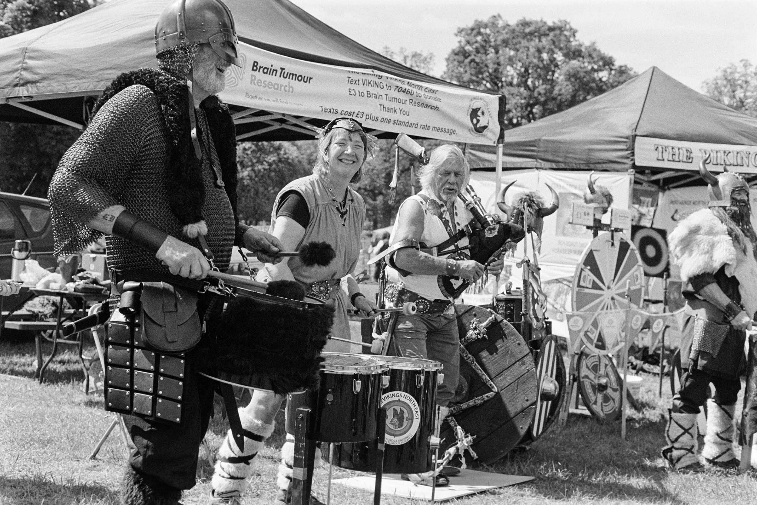 A group of people dressed in Viking costumes at an outdoor event, with tents and banners in the background, some playing musical instruments and others chatting.