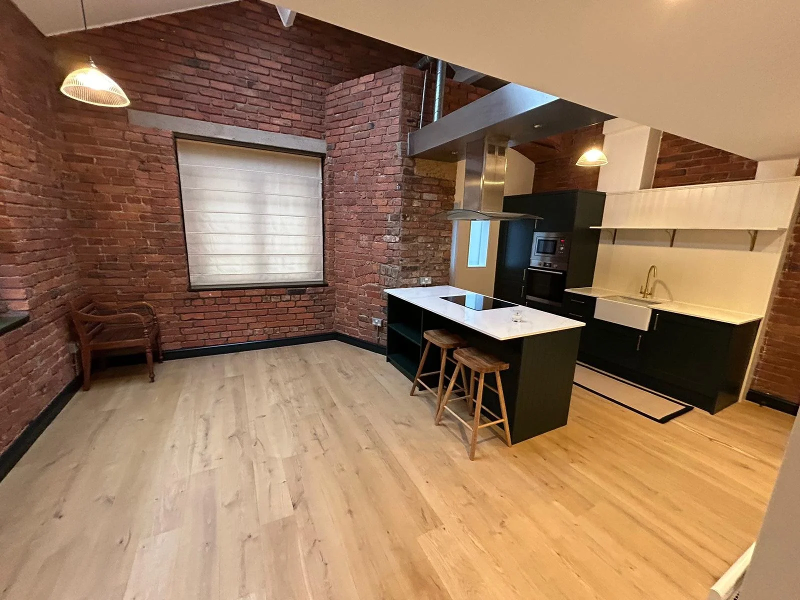 Modern kitchen with exposed brick walls, light wooden flooring, a white quartz worktop island with two stools, built-in black appliances, open shelving, a window with a shade, and pendant lighting.