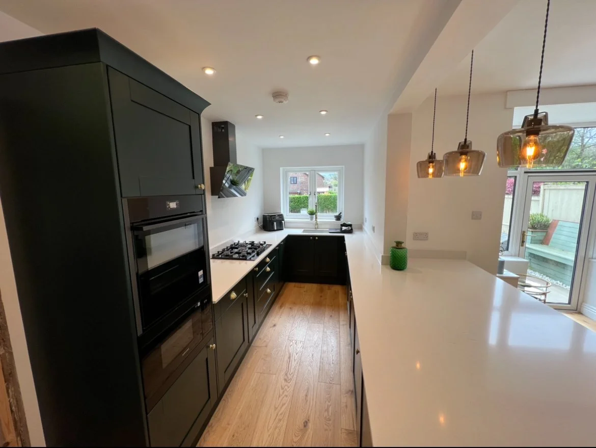 Modern kitchen with black cabinetry, white countertops, wooden floor, and pendant lights. There's a window above the sink, and a sliding door leading outside.