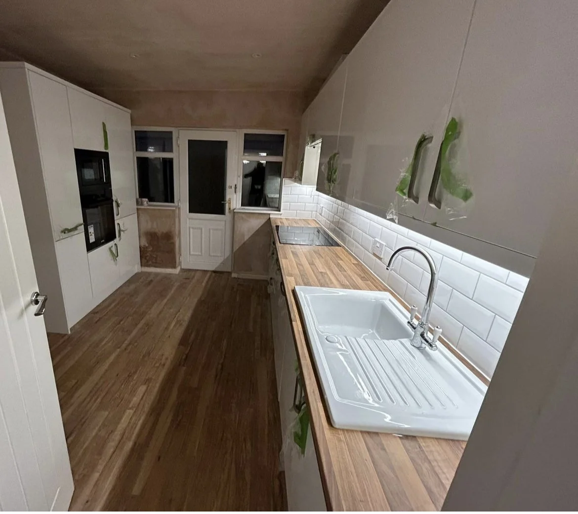 A modern kitchen with wooden countertops, white cabinetry, a white tiled backsplash, a sink, and appliances, with a door and window at the far end.