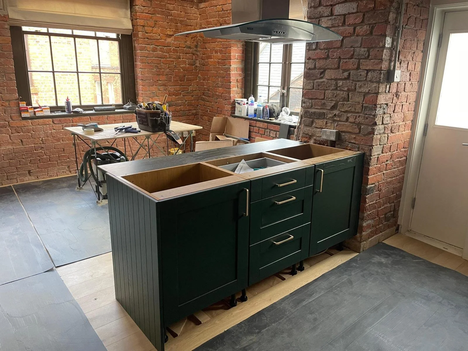 Kitchen under renovation with brick walls, a green kitchen island cabinet being installed, a window with tools and supplies on the window sill, and a work table with tools and a black tool bag in a room with black protective floor coverings.