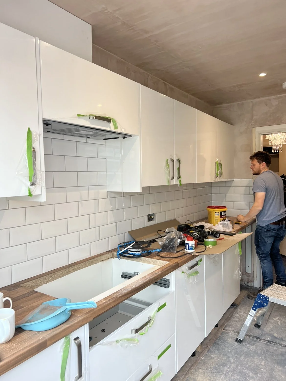 Kitchen under renovation with white cabinets, white subway tile backsplash, countertop with tools and supplies, a man working on the right side, and a step ladder.