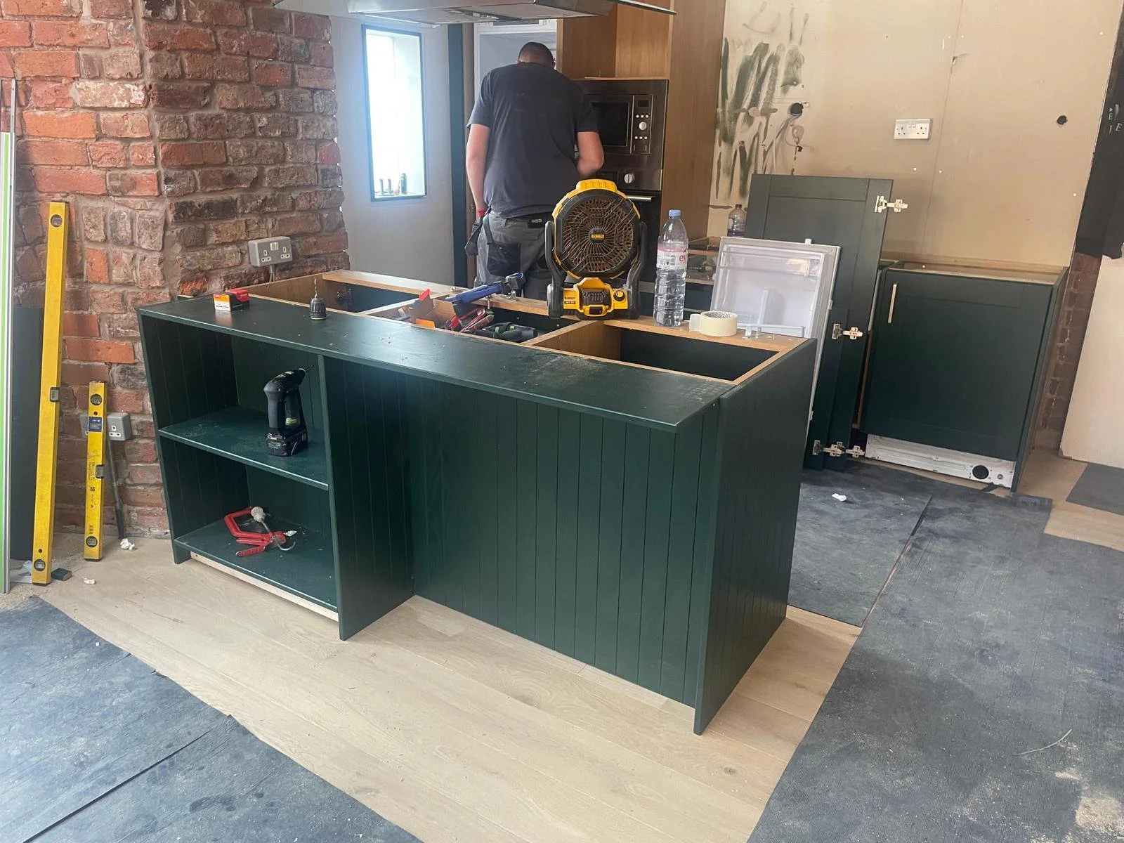Kitchen under construction with a green island in the foreground, tools on it, and a man working in the background near wall cabinets and an oven.